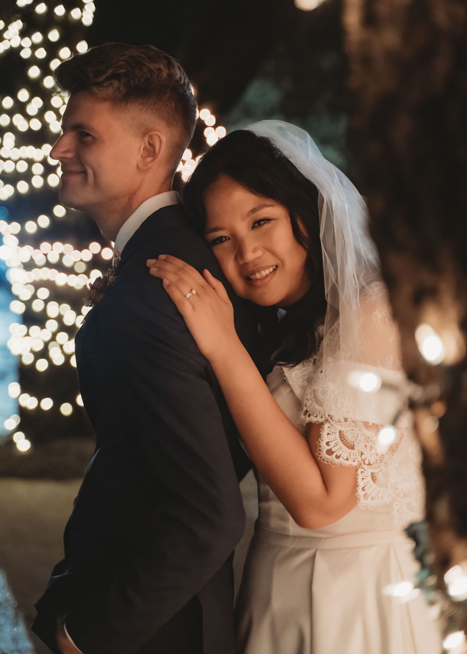 Bride and groom embrace outdoors at night with warm fairy lights bokeh background at romantic wedding celebration.