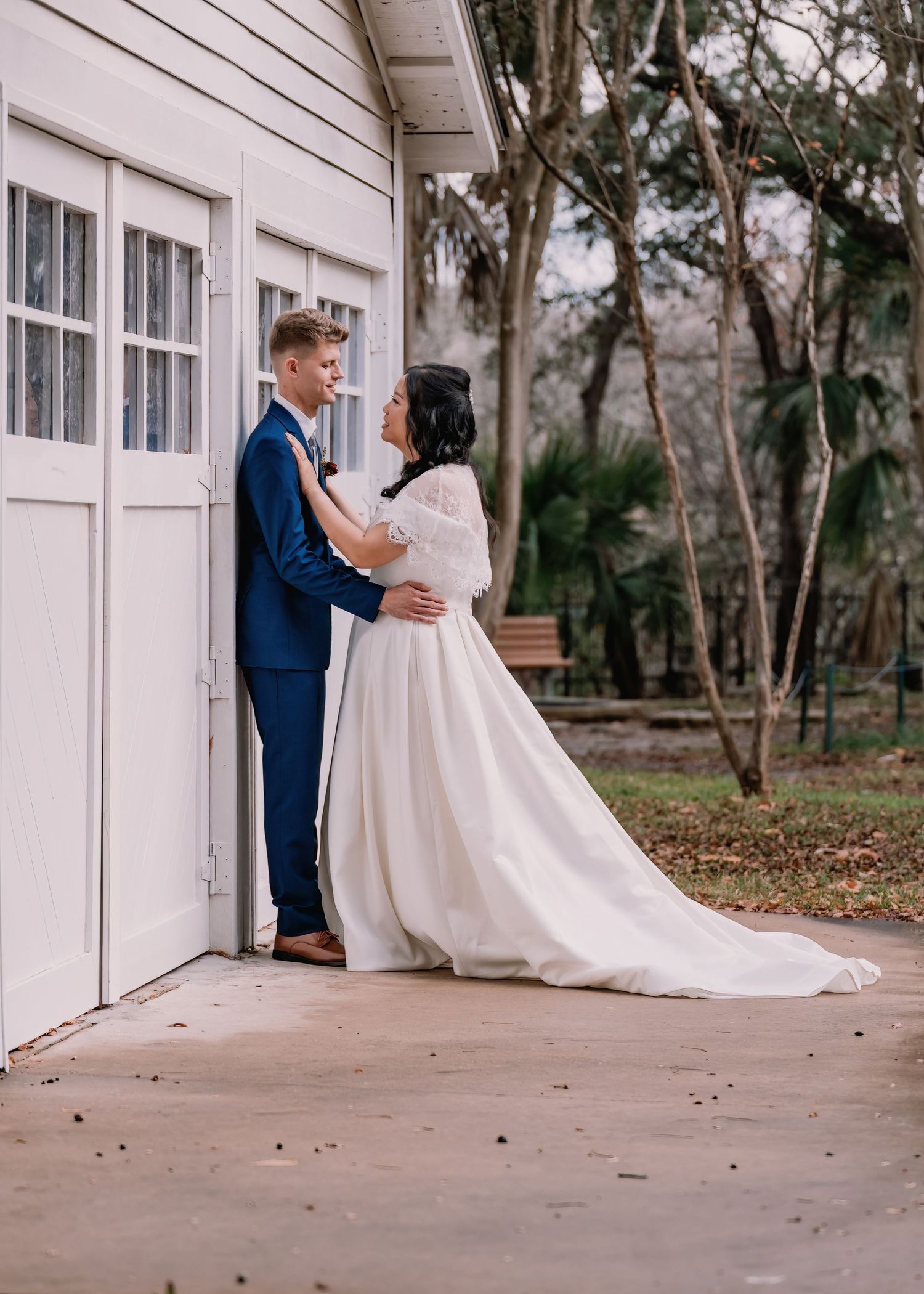 A bride in a white gown and groom in a navy suit share a romantic moment by white barn doors surrounded by trees.