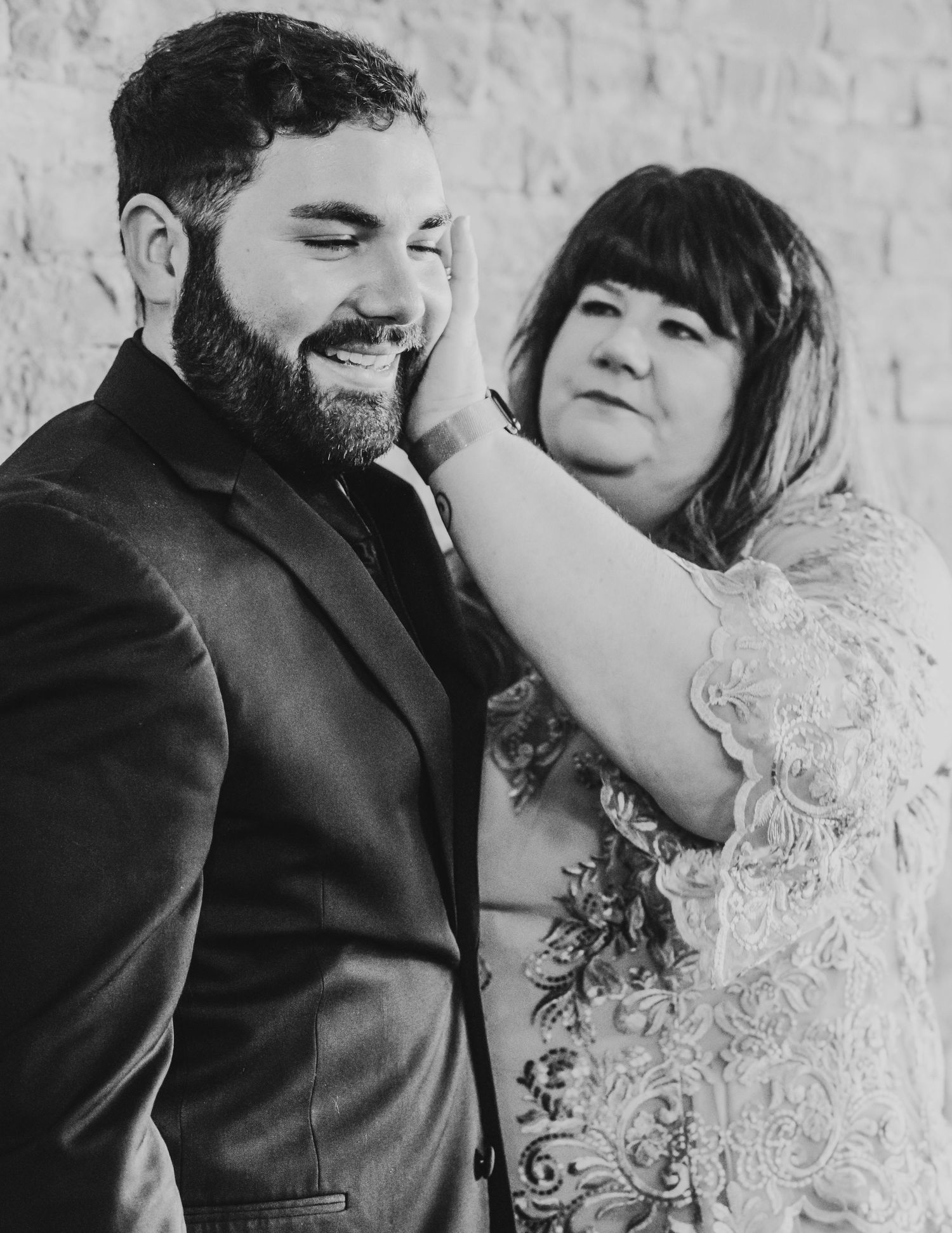 Black and white photo of a bearded groom smiling as a woman in lace dress tenderly touches his face.
