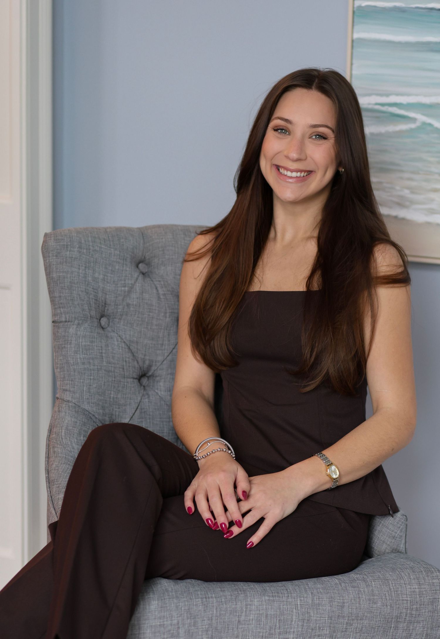 Woman in black dress relaxes on gray tufted sofa with coastal artwork and natural lighting.
