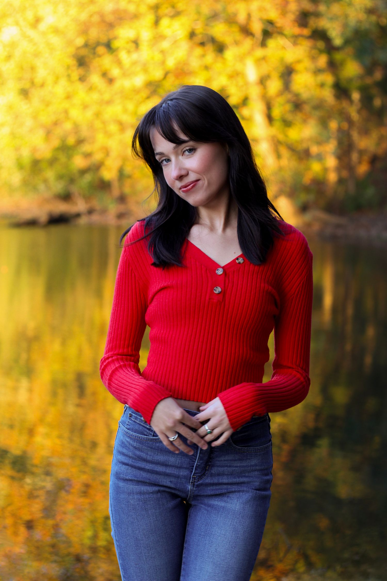 Person poses by lake wearing red sweater against yellow fall foliage backdrop.