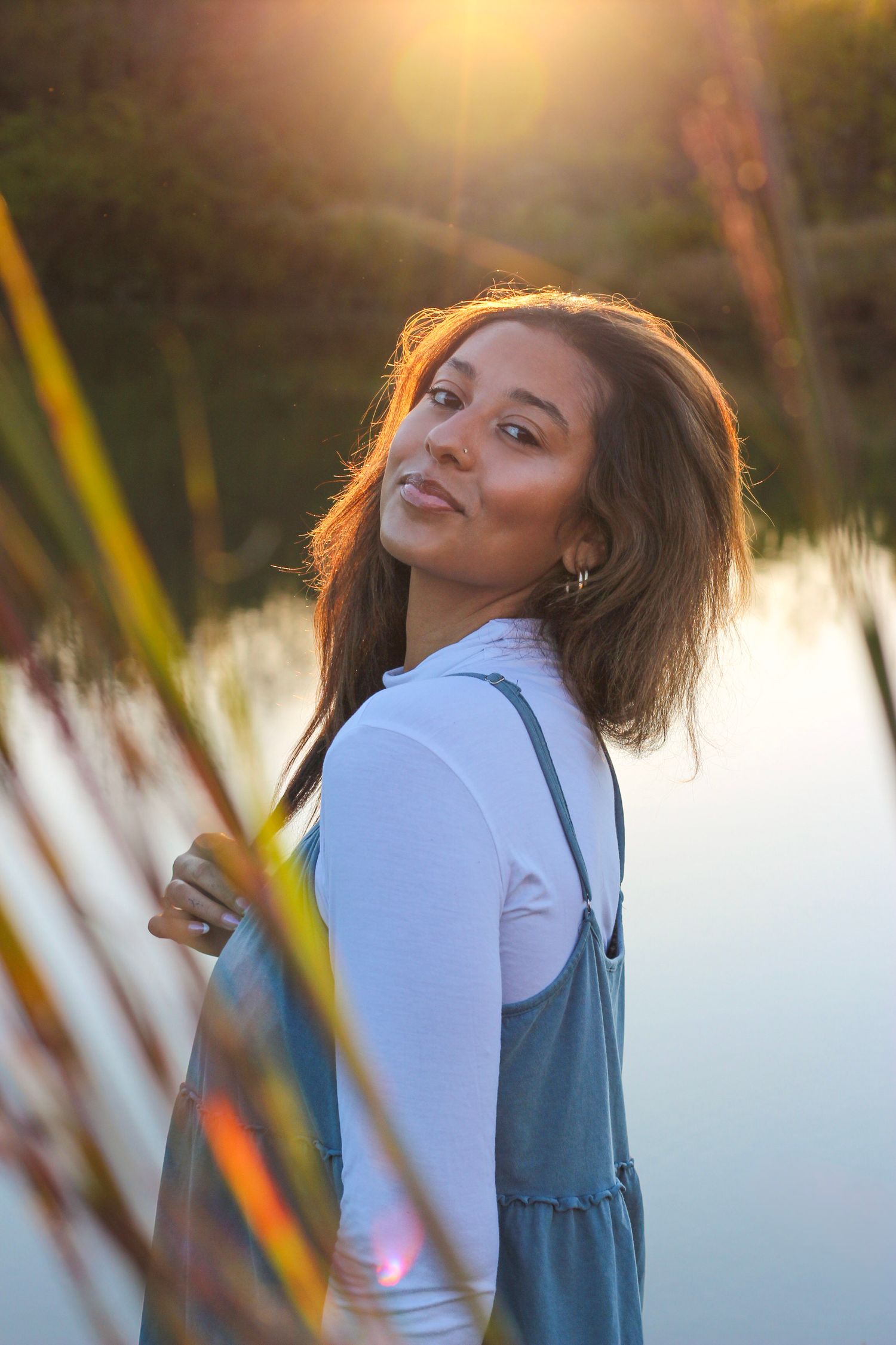 Woman in white shirt and blue overalls poses playfully against golden sunset by a lake.