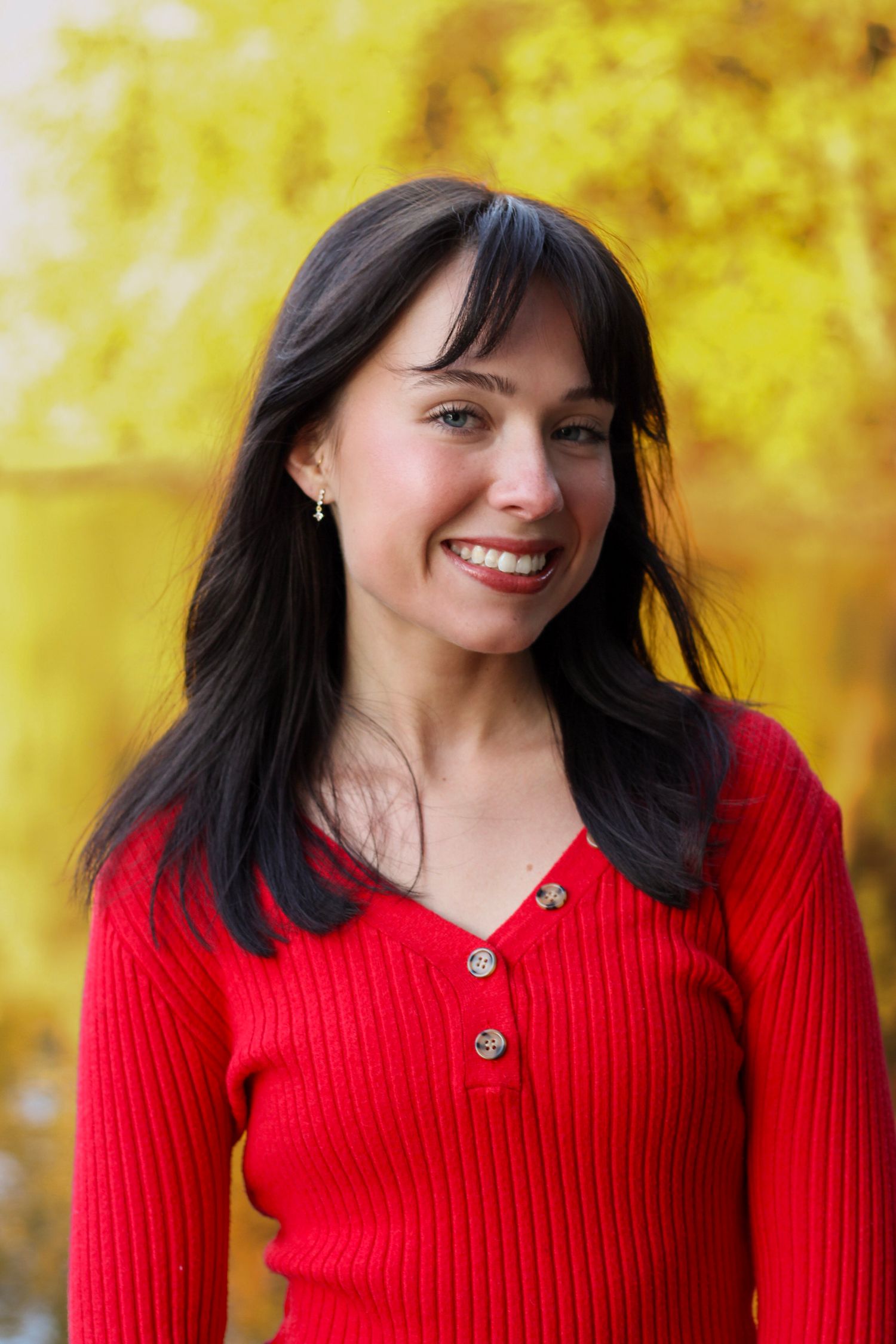 Person wearing red knit top poses outdoors with yellow autumn trees in background.