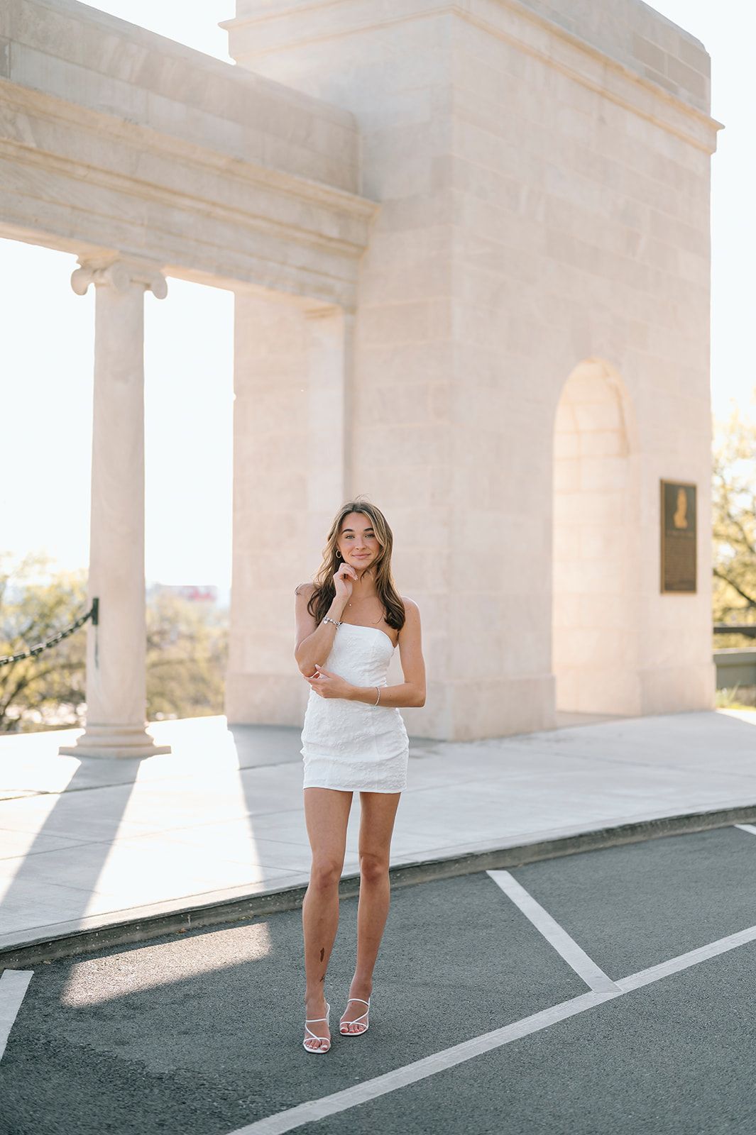 Person in white mini dress standing by architectural columns on sunny day.