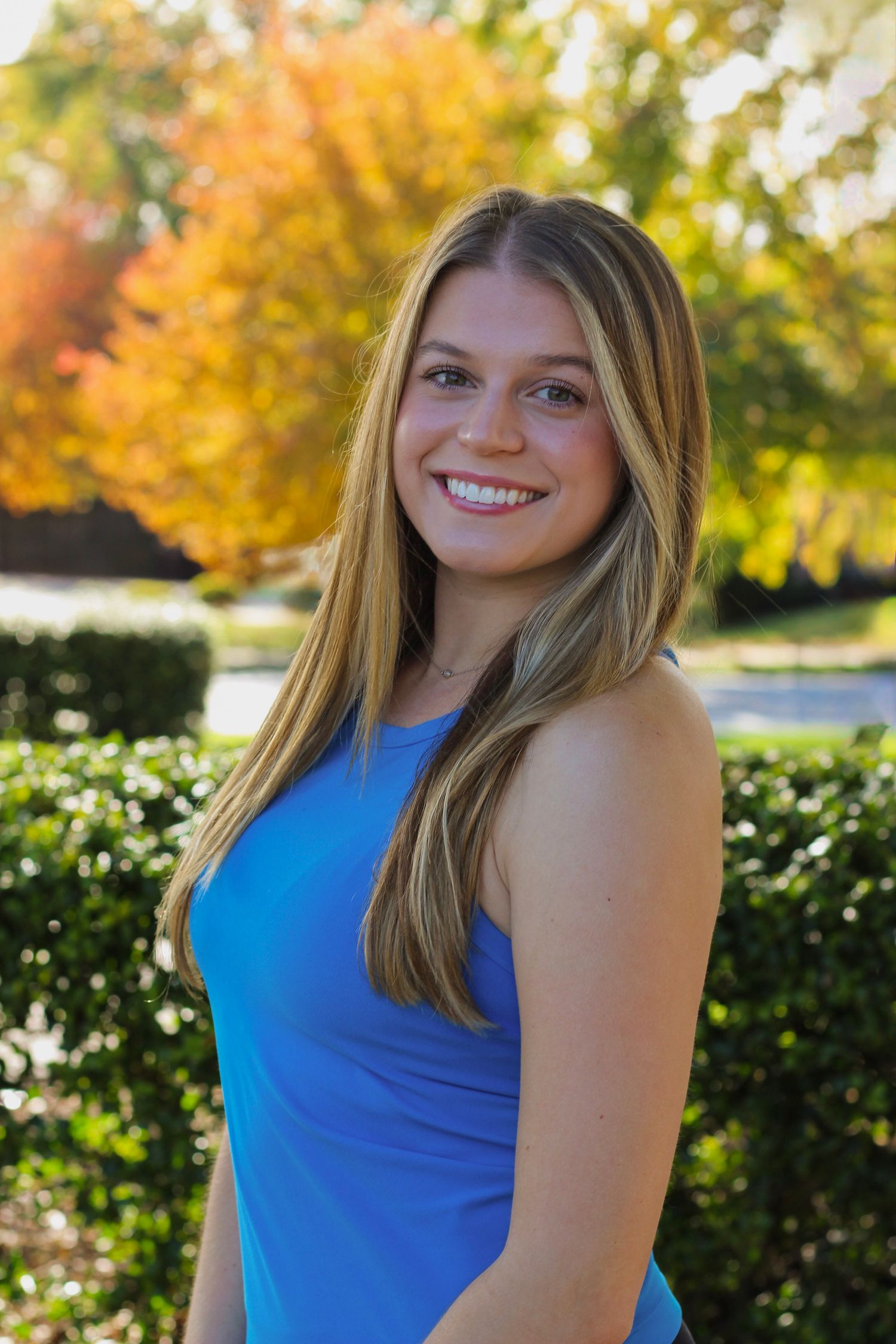 Portrait series in blue tank top against autumn trees and garden landscaping.