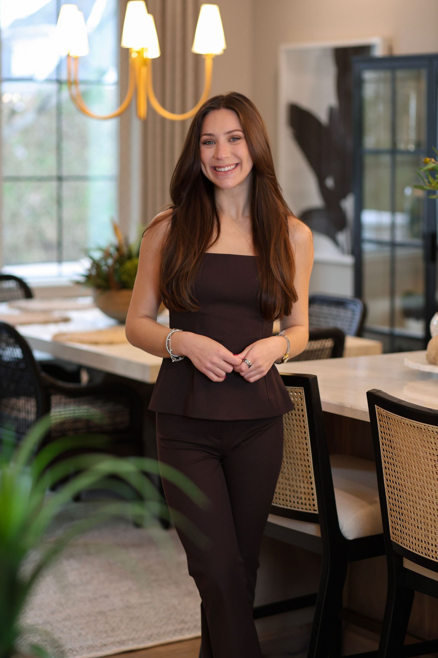 Woman in black dress sits casually at dining table with cane chairs and warm lighting.