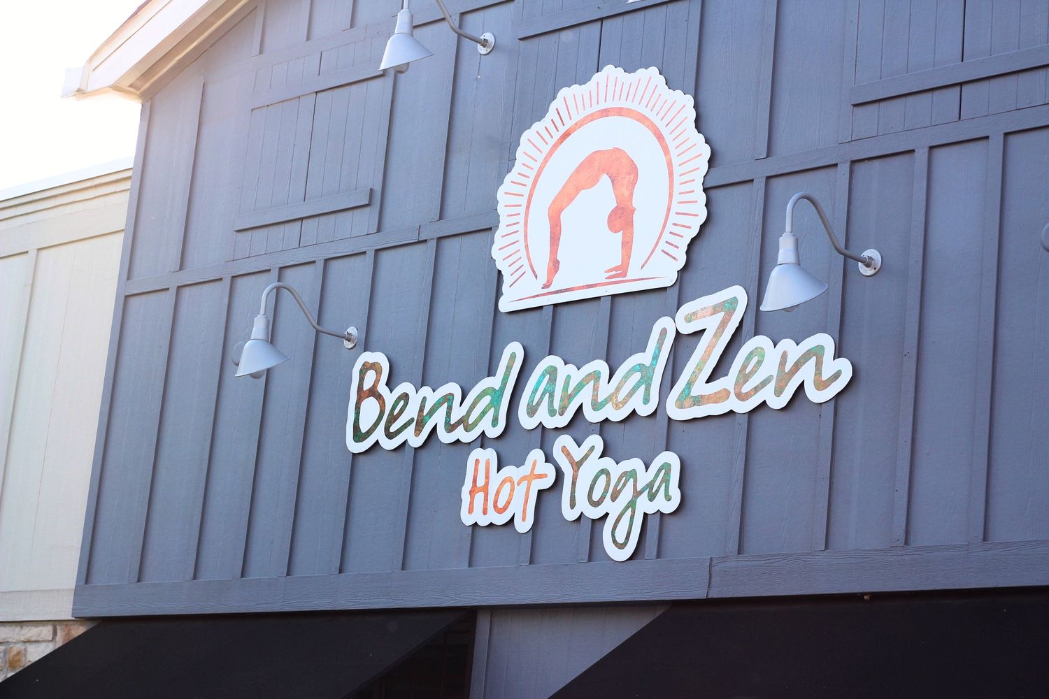 Exterior sign for 'Bend and Zen Hot Yoga' studio with logo on gray building.