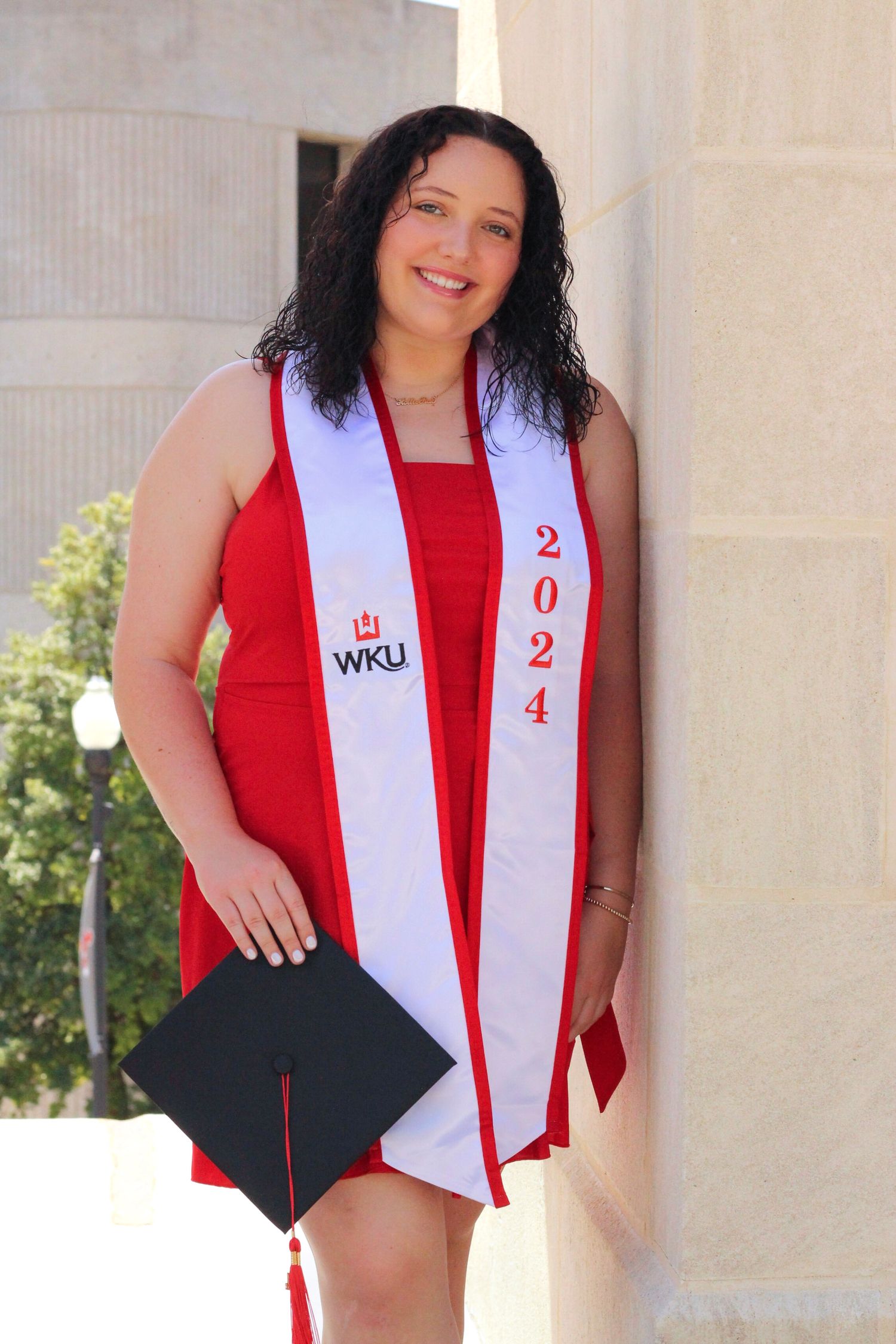 Graduate in red dress and white WKU sash poses with graduation cap against classical columns.