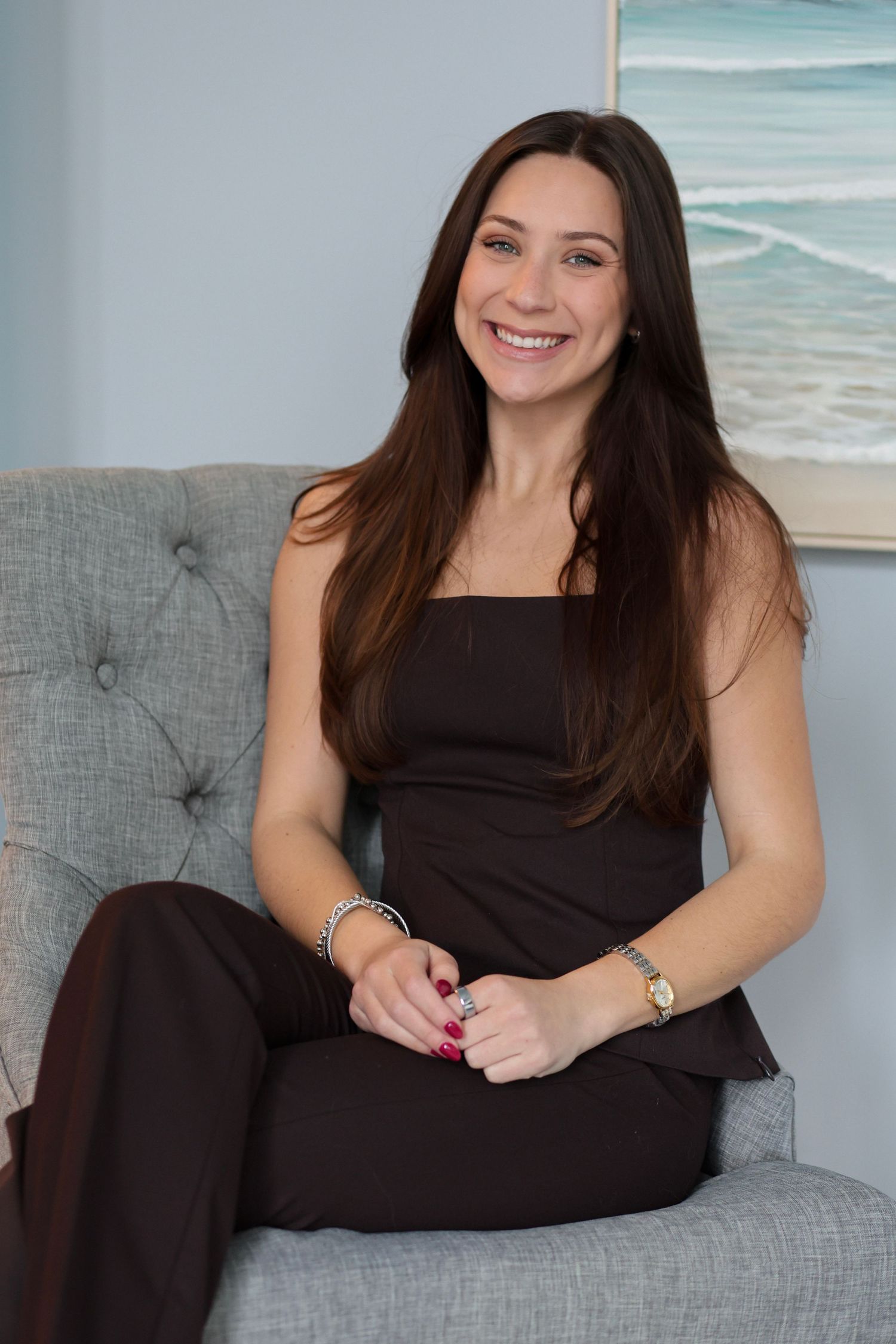 Woman in black dress relaxes on gray tufted couch with coastal artwork in background.