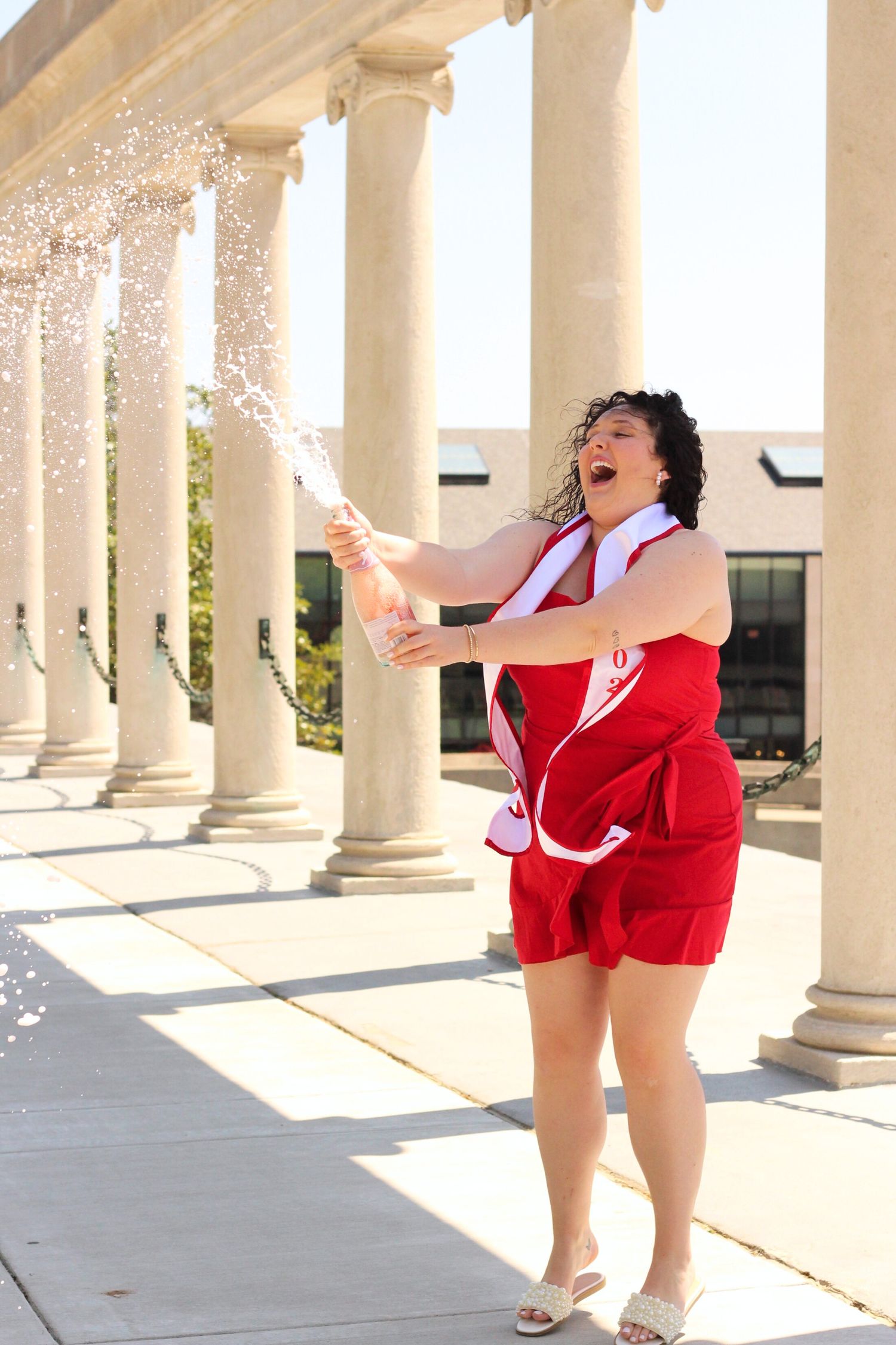 Graduate in red dress celebrates joyfully under white stone colonnade archway.