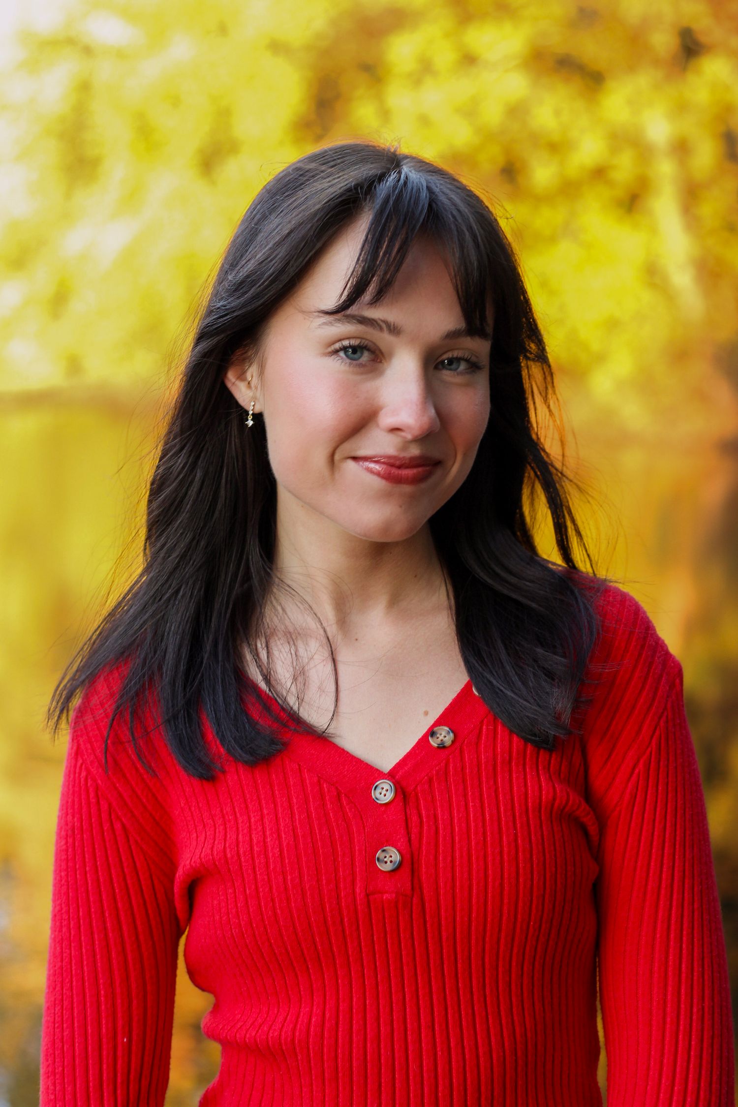 Person in bright red ribbed sweater smiles against vibrant autumn foliage background.