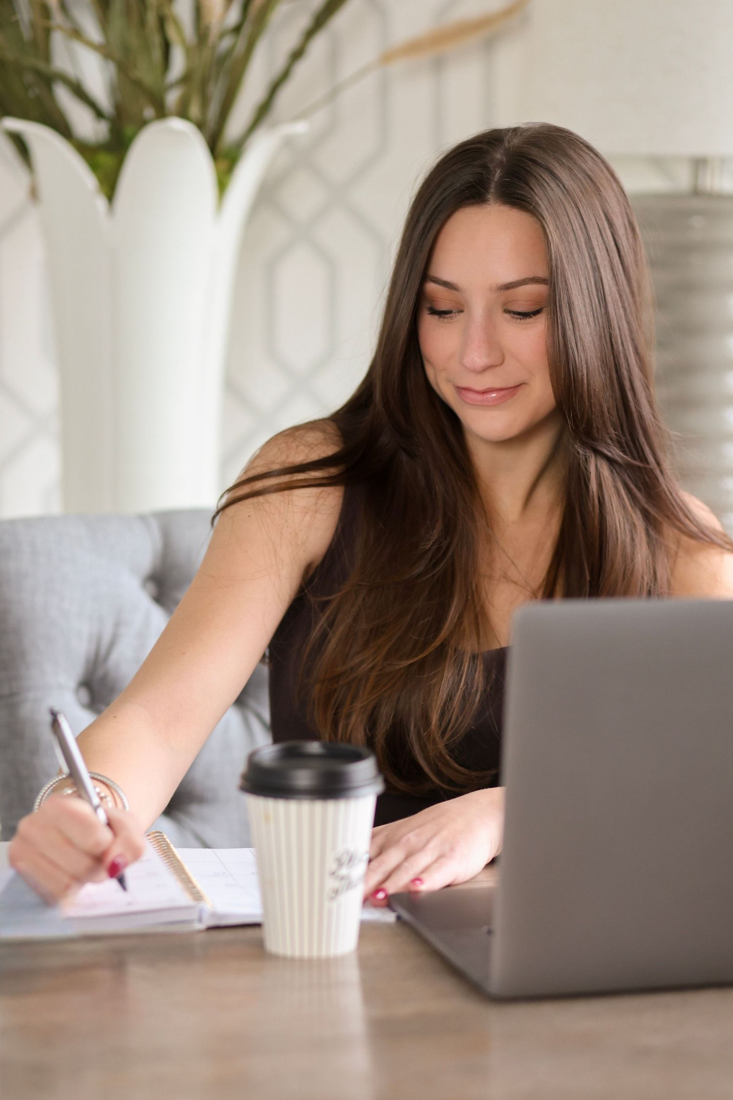 Person writing in notebook at wooden desk with laptop and coffee cup in bright modern workspace.