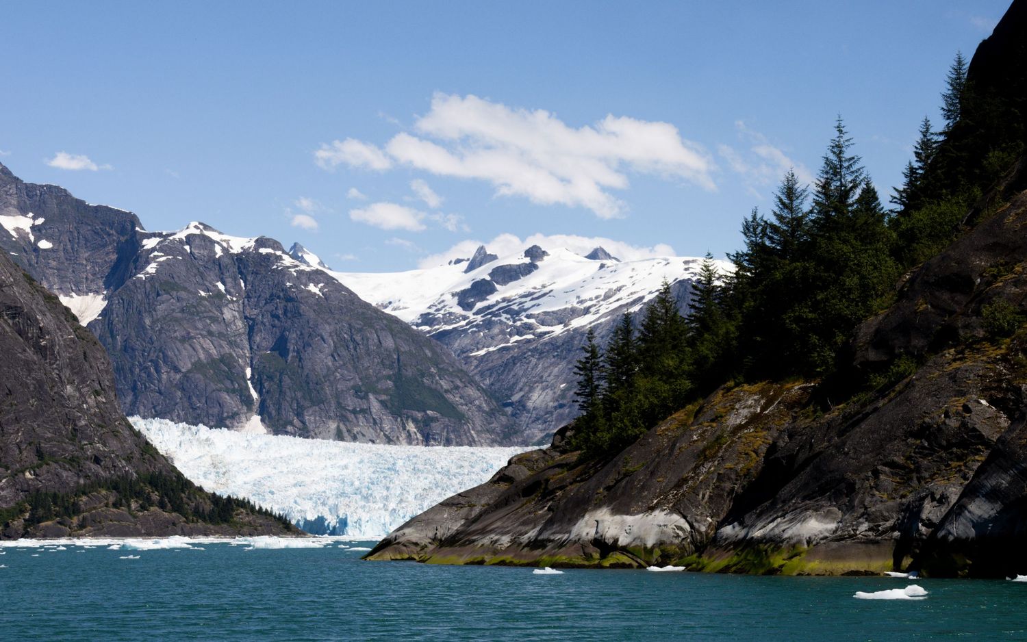 A scenic panorama of snow-capped mountains and glaciers reflecting in turquoise waters of an Alaskan fjord.