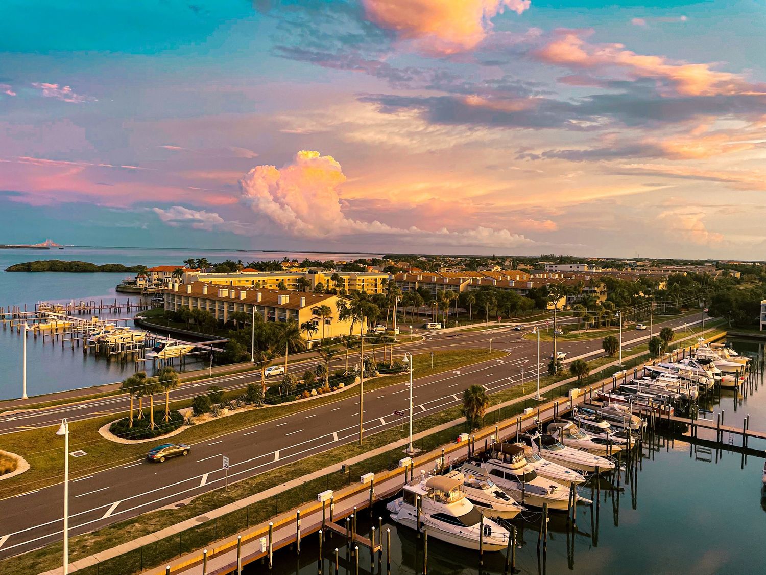 Panoramic sunset view of a coastal marina with boats docked along the waterfront and palm trees lining the shore.