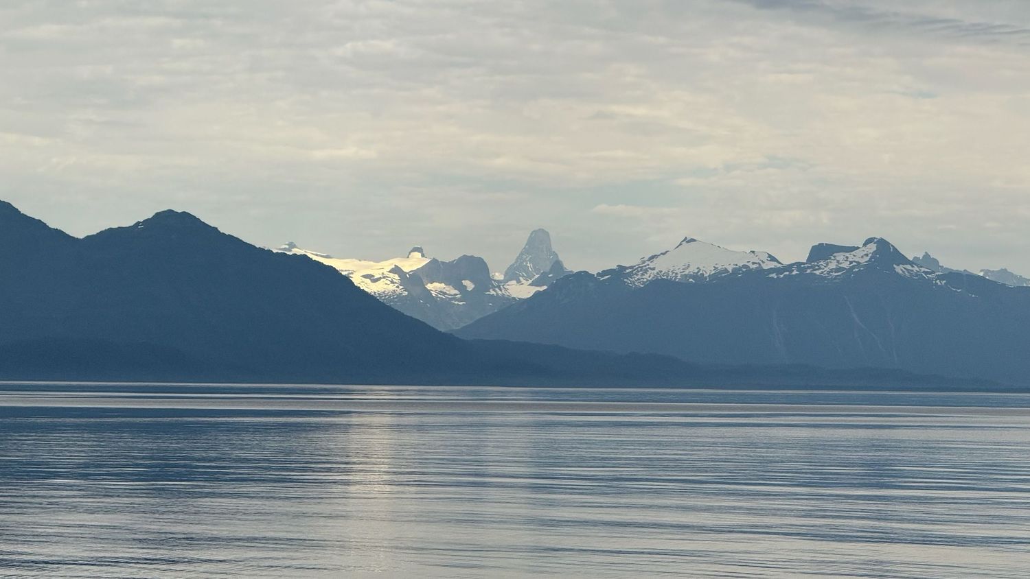 Panoramic view of snow-capped mountains reflecting in calm waters of an Alaskan fjord at dusk.