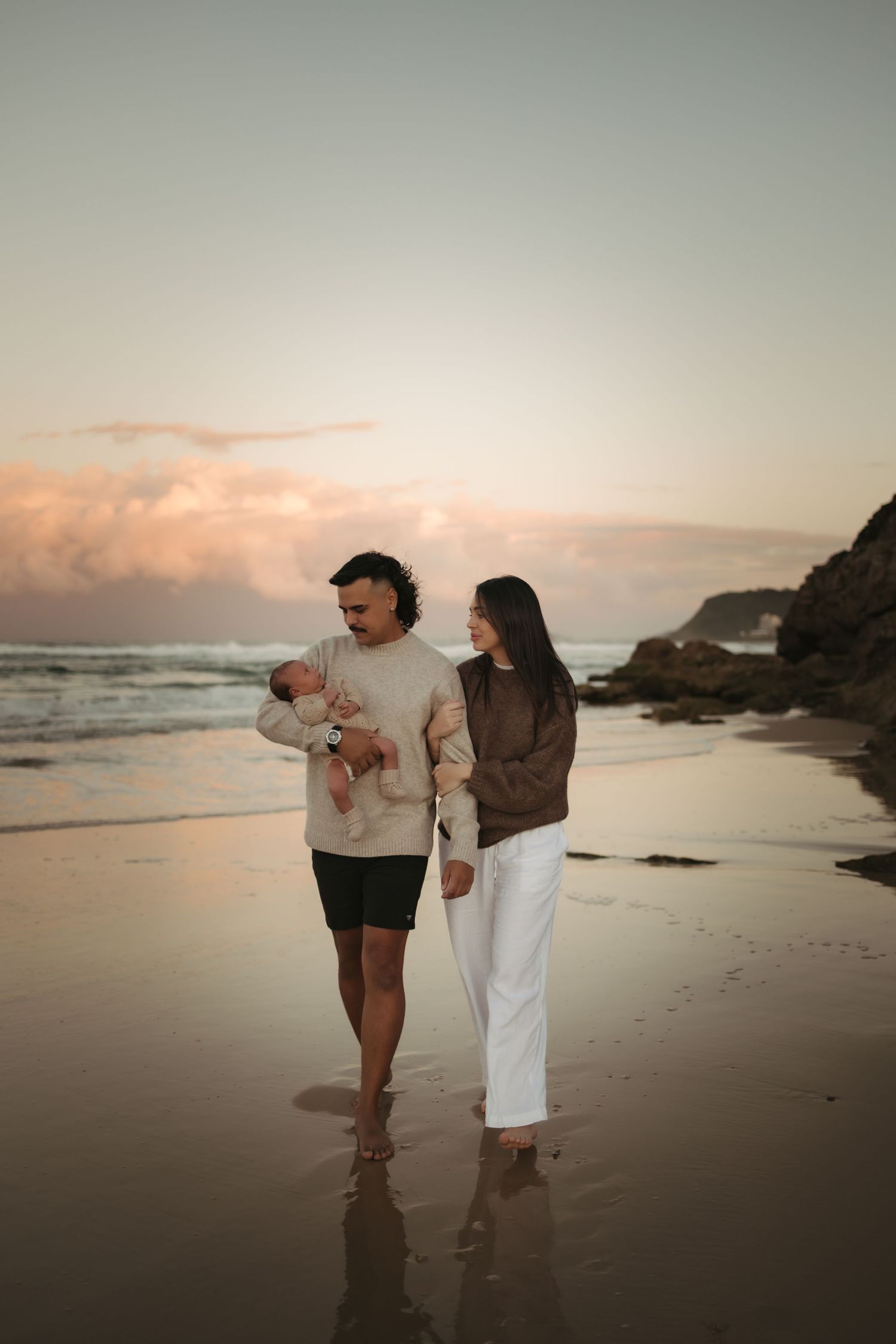 A couple walks along a misty beach at sunset with rocky cliffs in the background creating a romantic silhouette.
