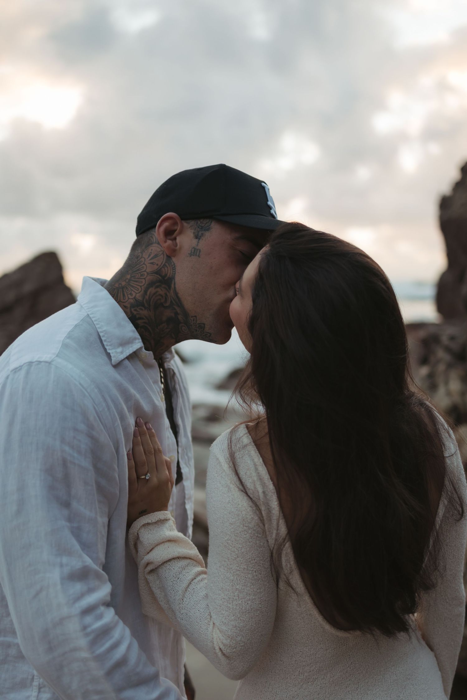 A romantic couple sharing a tender kiss at sunset against a cloudy sky and rocky backdrop.