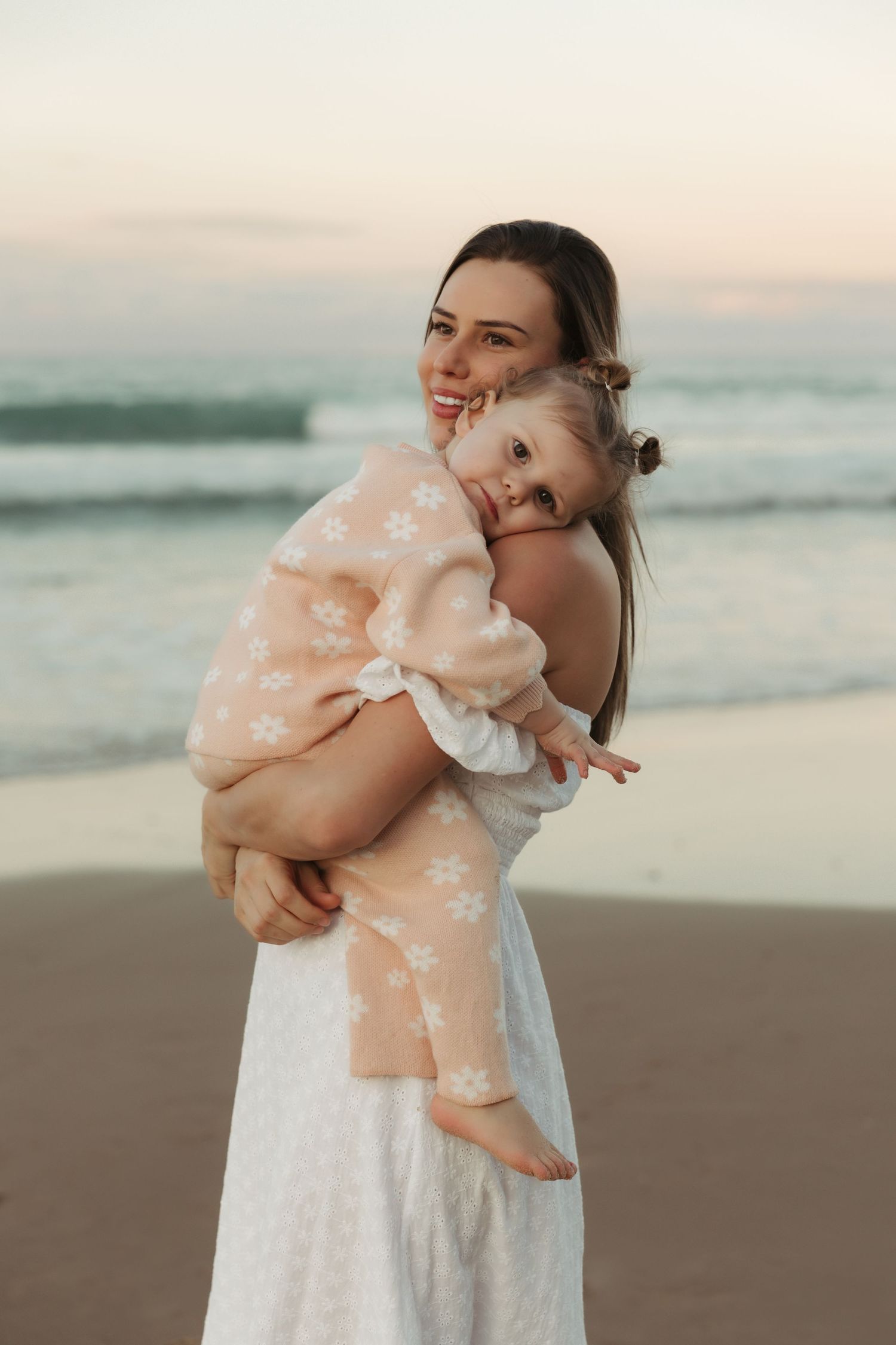 A tender beach sunset moment with a white-dressed woman holding a child in peachy attire on the shoreline.