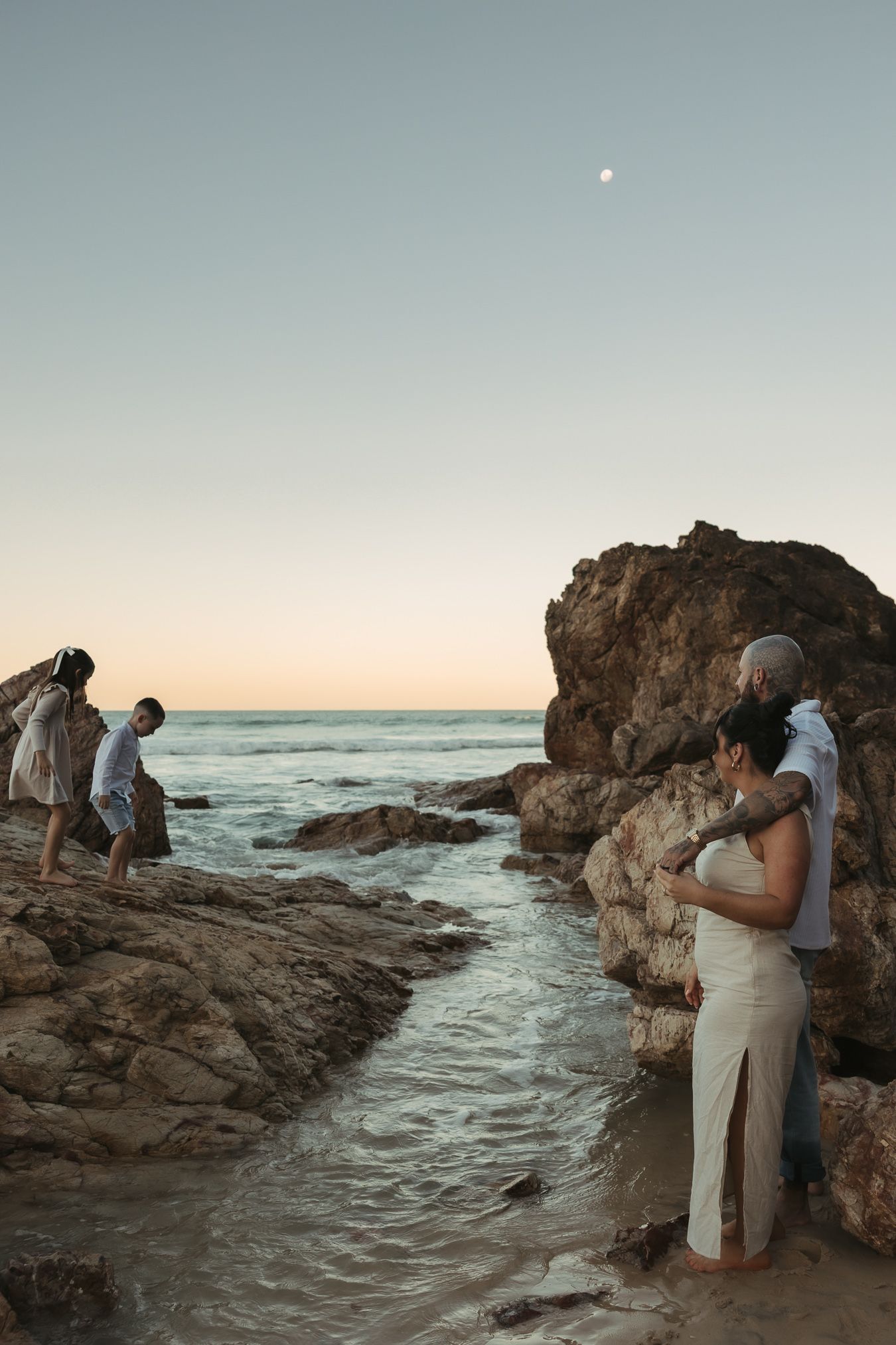 A couple shares a romantic moment at sunset by a rocky ocean arch formation on the beach.