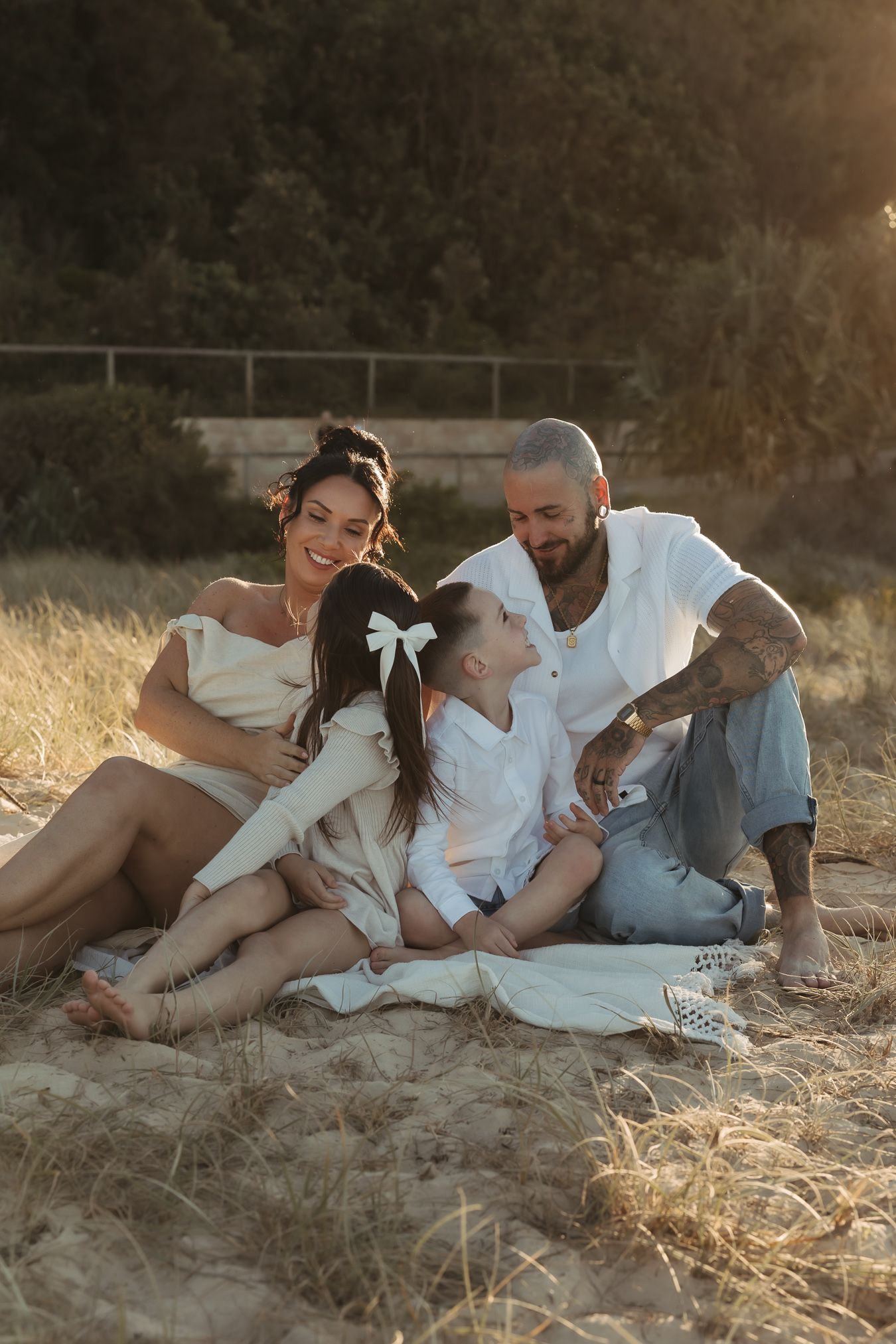 Family lounging together in casual white outfits on golden grass hillside.