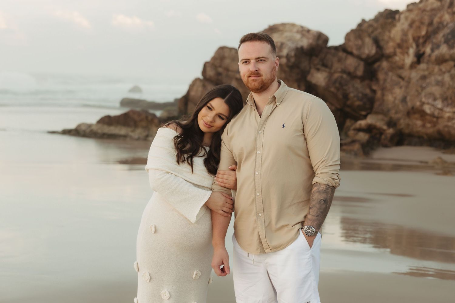 A couple in light-colored attire poses lovingly together on a scenic rocky beach at sunset.
