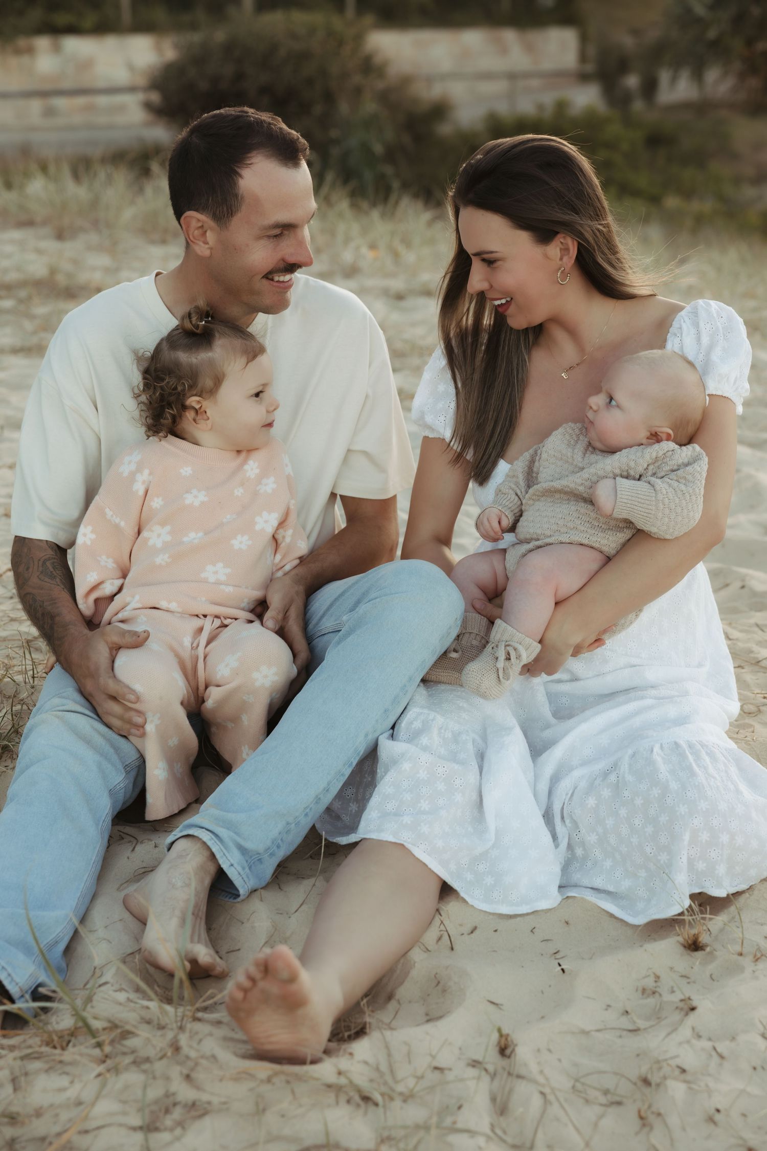 A couple sits together on a sandy beach wearing light colored clothing while sharing intimate moments.