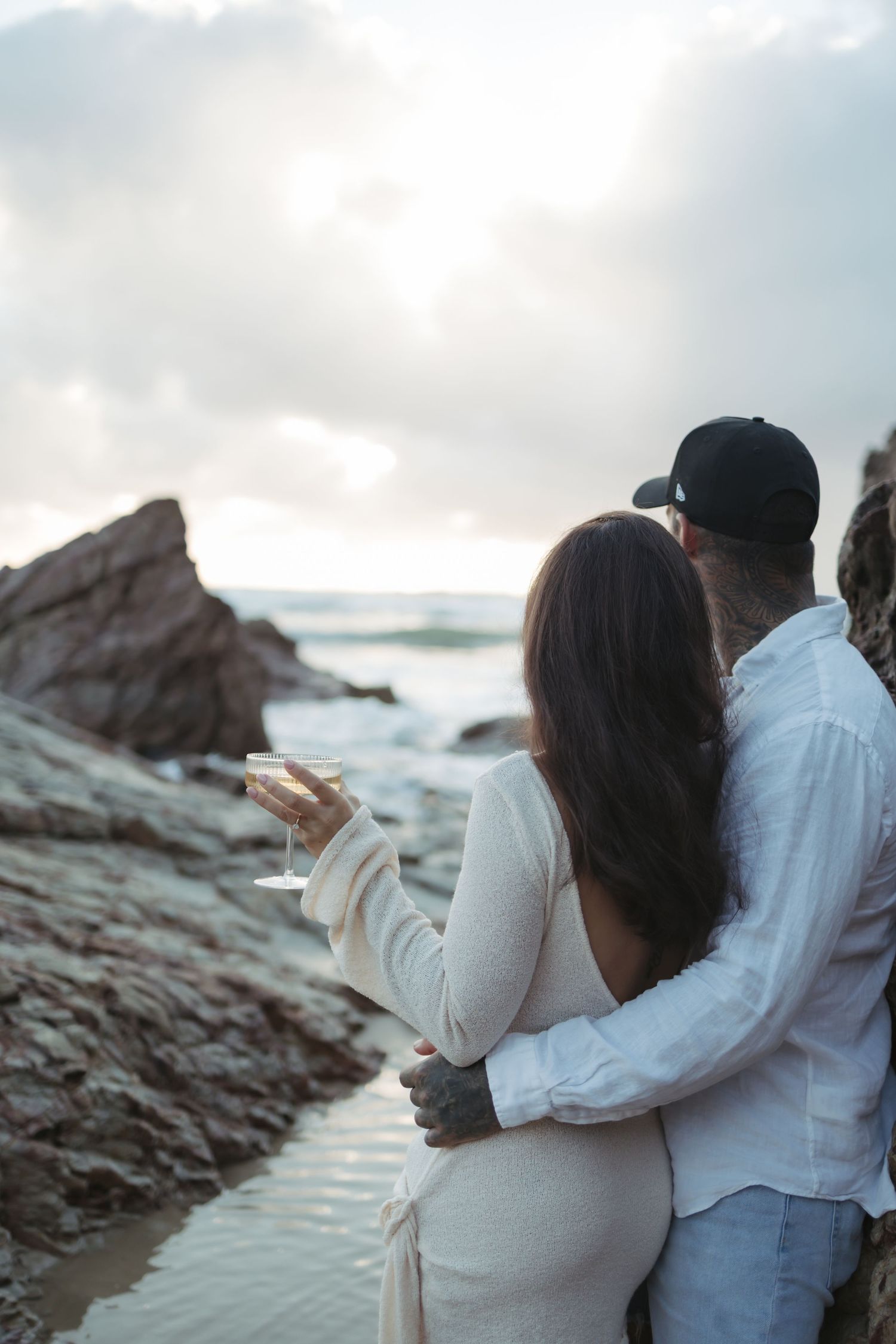 A couple embraces while sitting on rocky coastal cliffs at sunset as ocean waves crash in the background.