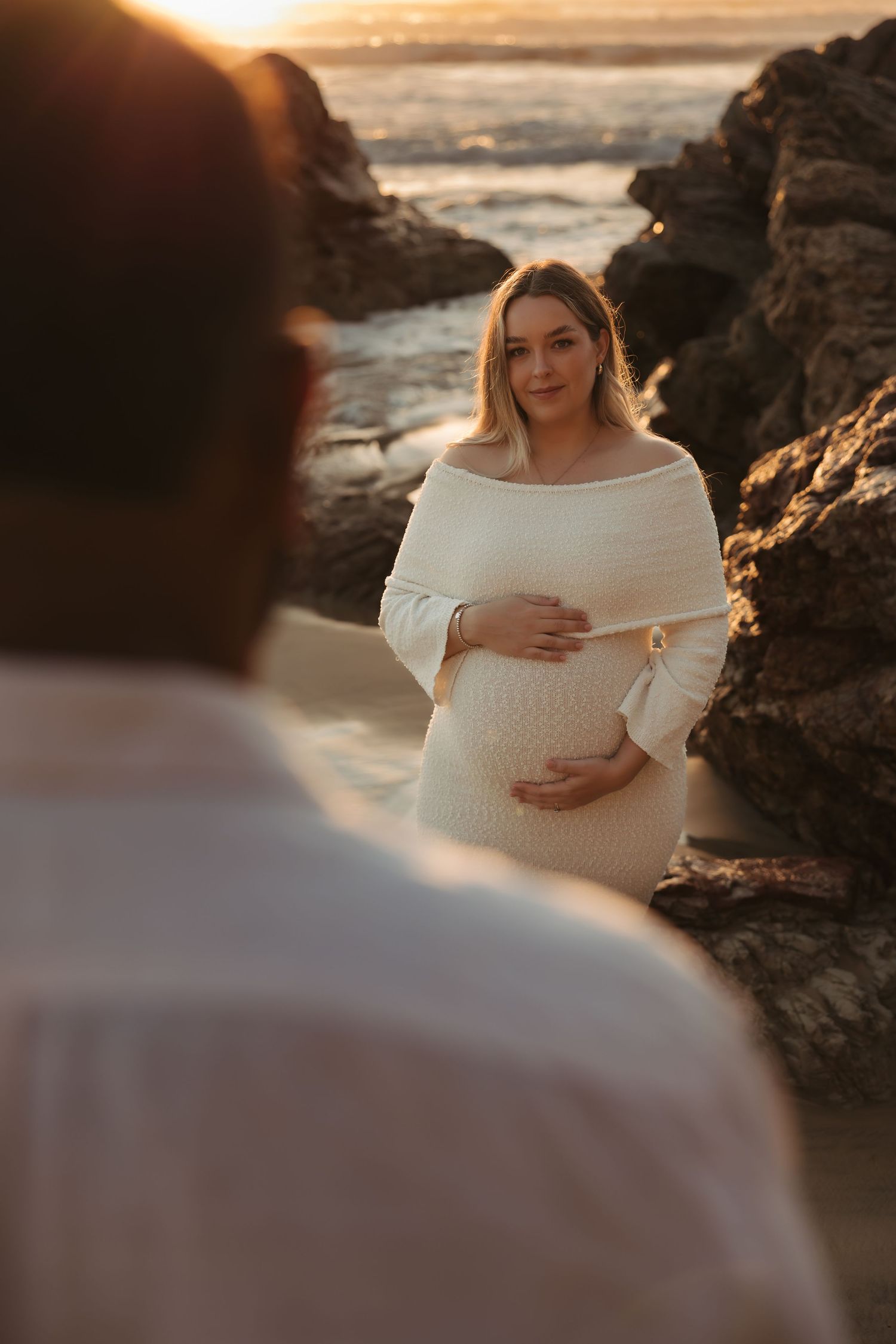 A maternity photoshoot at sunset on a rocky beach with warm golden light filtering through the coastal cliffs.