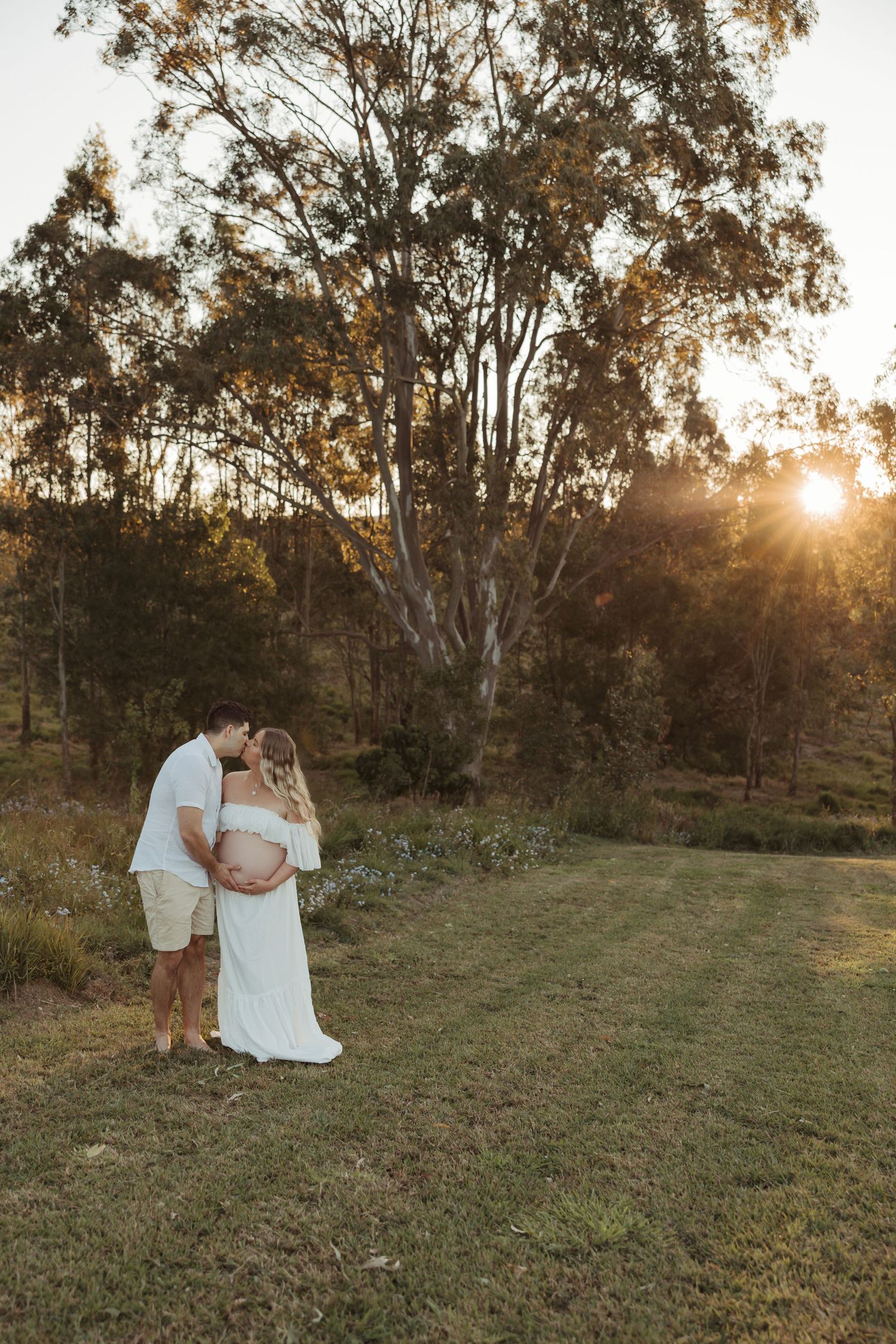 Romantic maternity photoshoot in a grassy field at sunset with warm golden light filtering through trees.