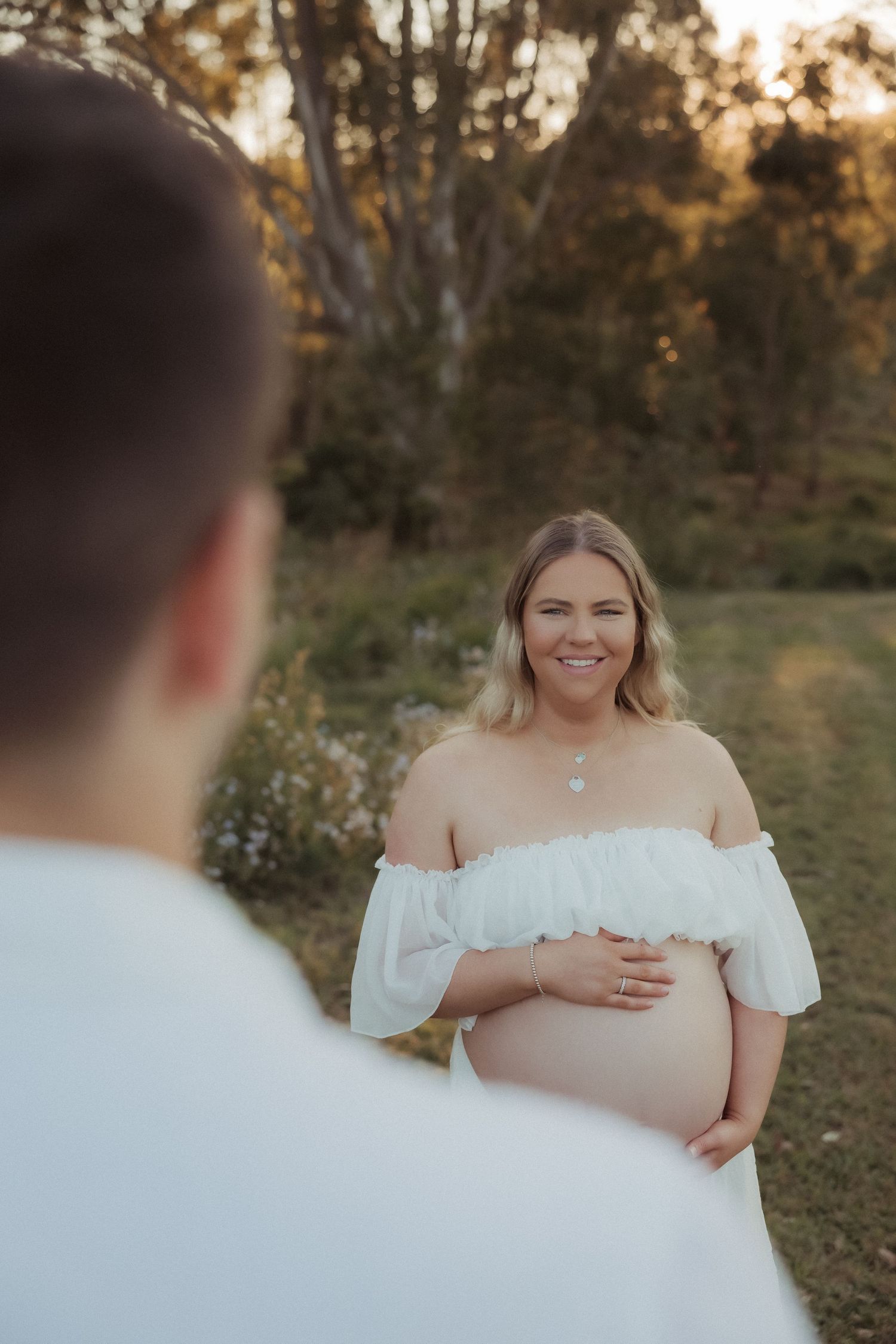 Expectant mother in white off-shoulder dress poses outdoors during golden hour maternity photoshoot.