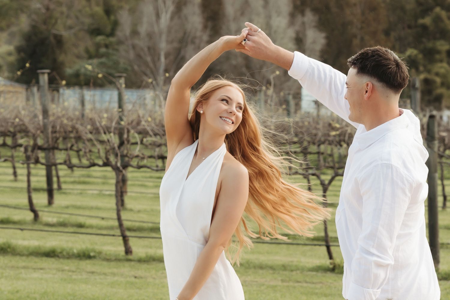 A couple in white outfits dances playfully together in a scenic outdoor orchard setting during golden hour.
