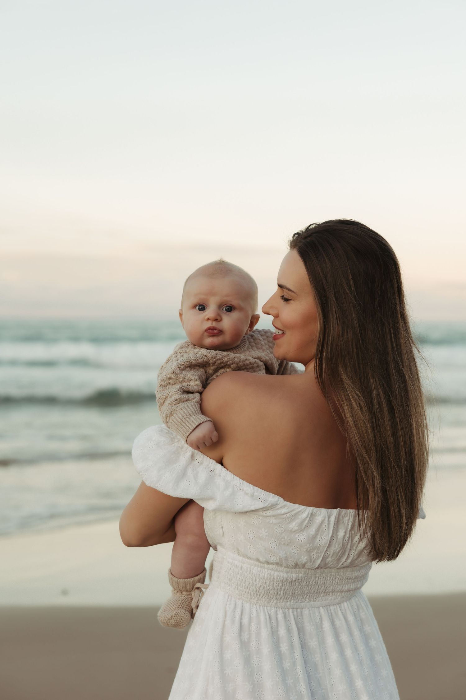 A tender moment between parent and baby in white clothing on a peaceful beach at sunset.