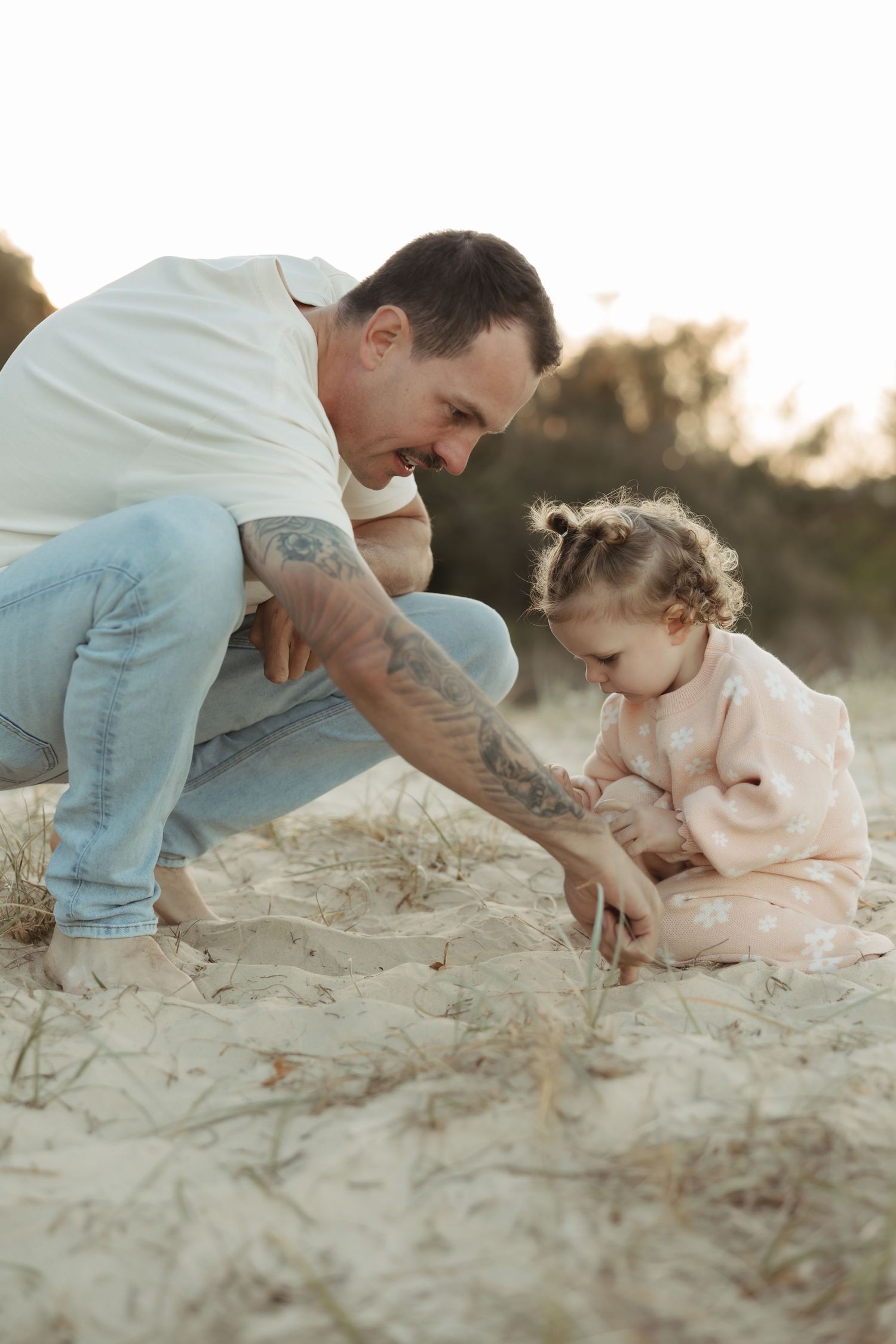 A parent and child crouch together on sandy ground during golden hour, sharing a tender moment of discovery.