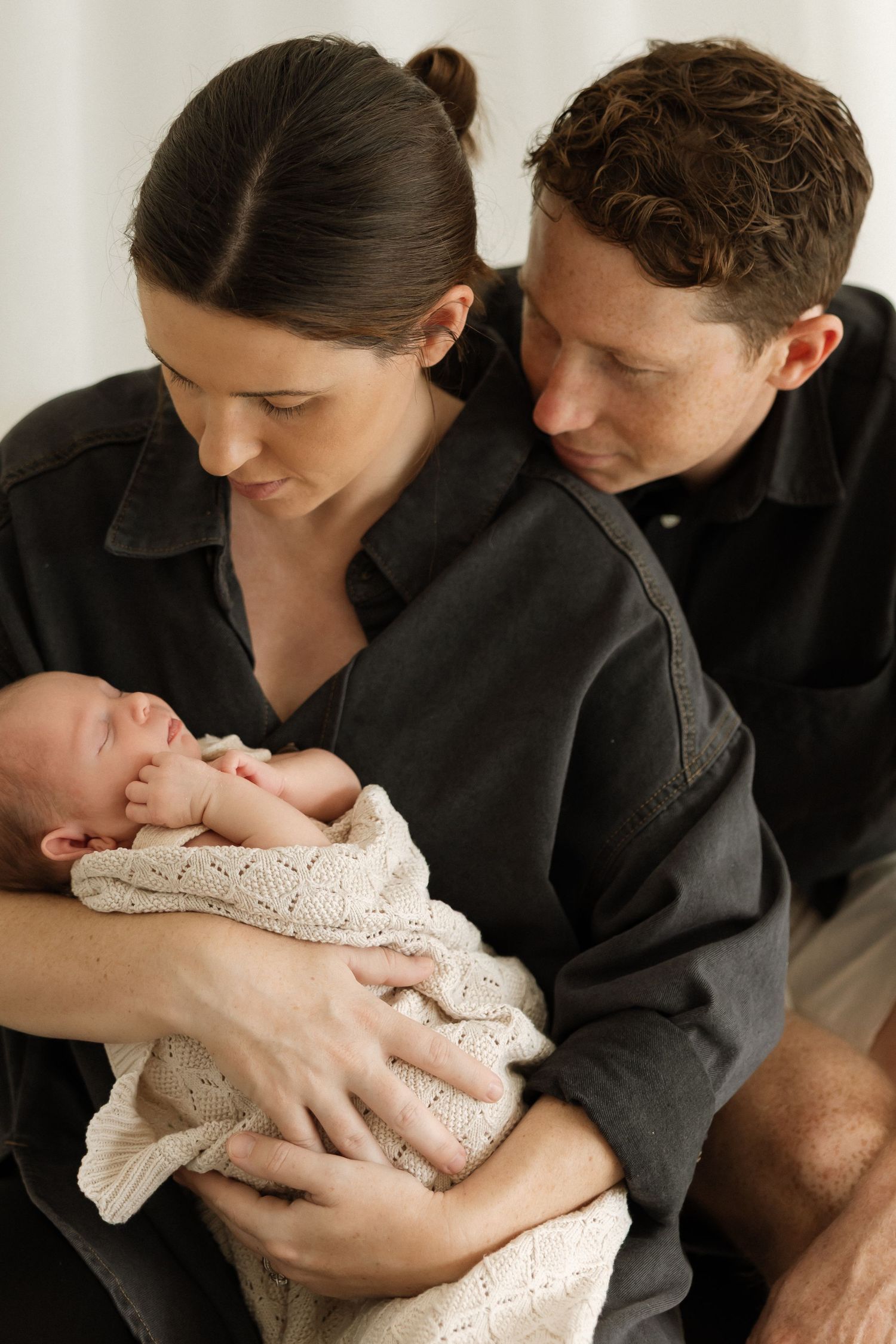 A tender family moment as parents in black shirts gaze lovingly at their newborn baby wrapped in a knit blanket.
