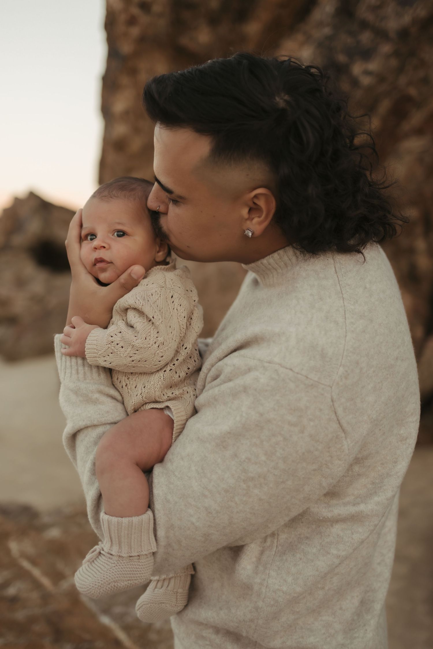 A tender moment captured in warm tones as a parent embraces and kisses their baby wearing a cream knit sweater.