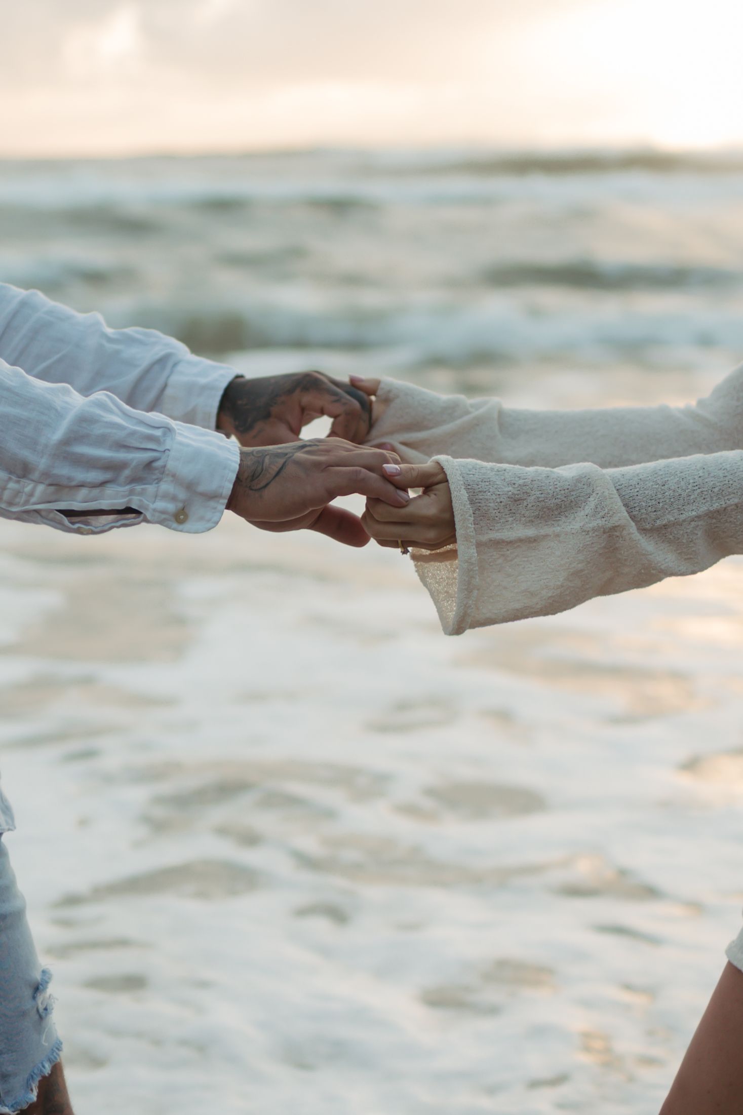 Two hands reach towards each other against a blurred beach background with soft sunlight.