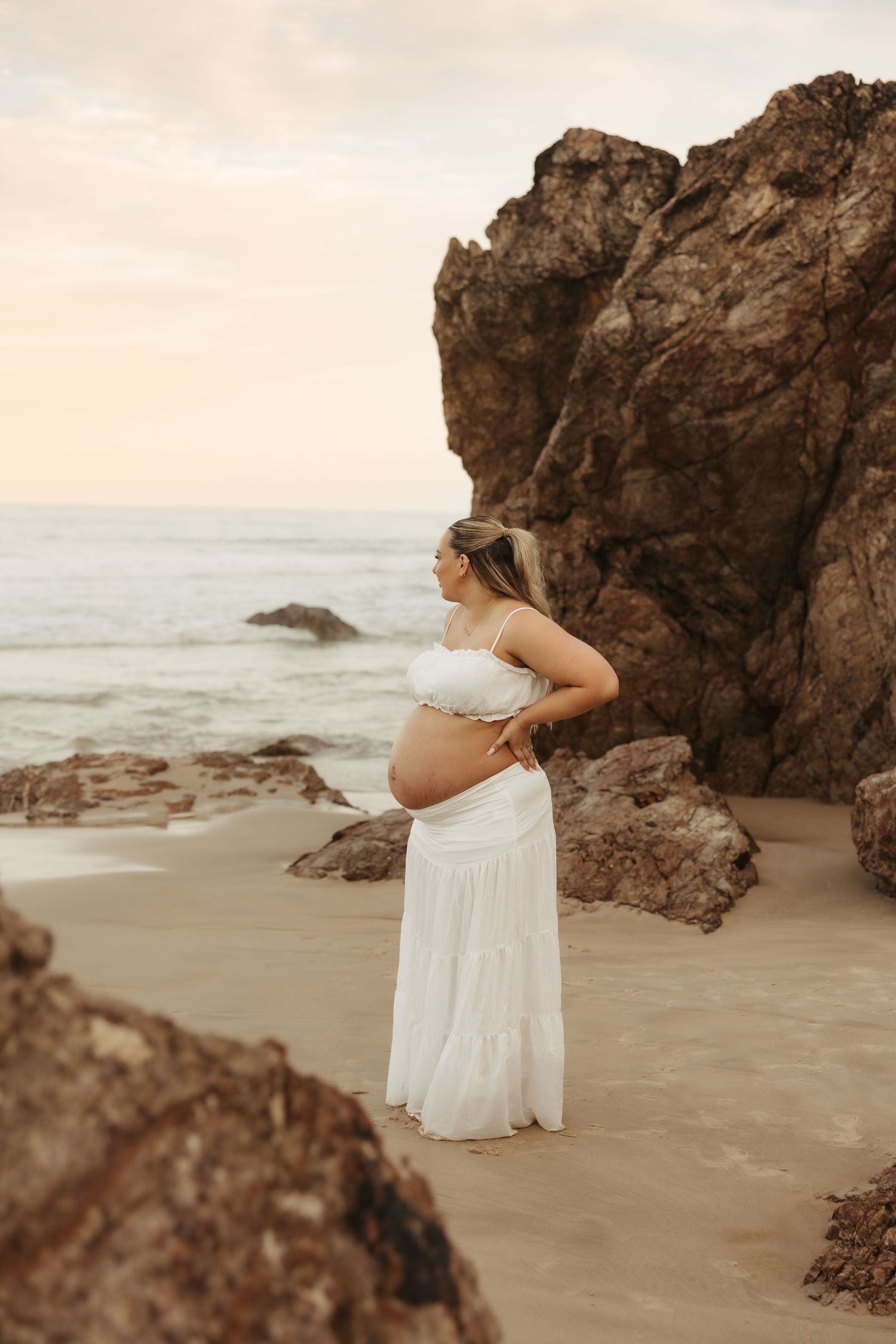 A series of maternity photos on a beach with rocky outcrops showing a person in a flowing white dress at sunset.
