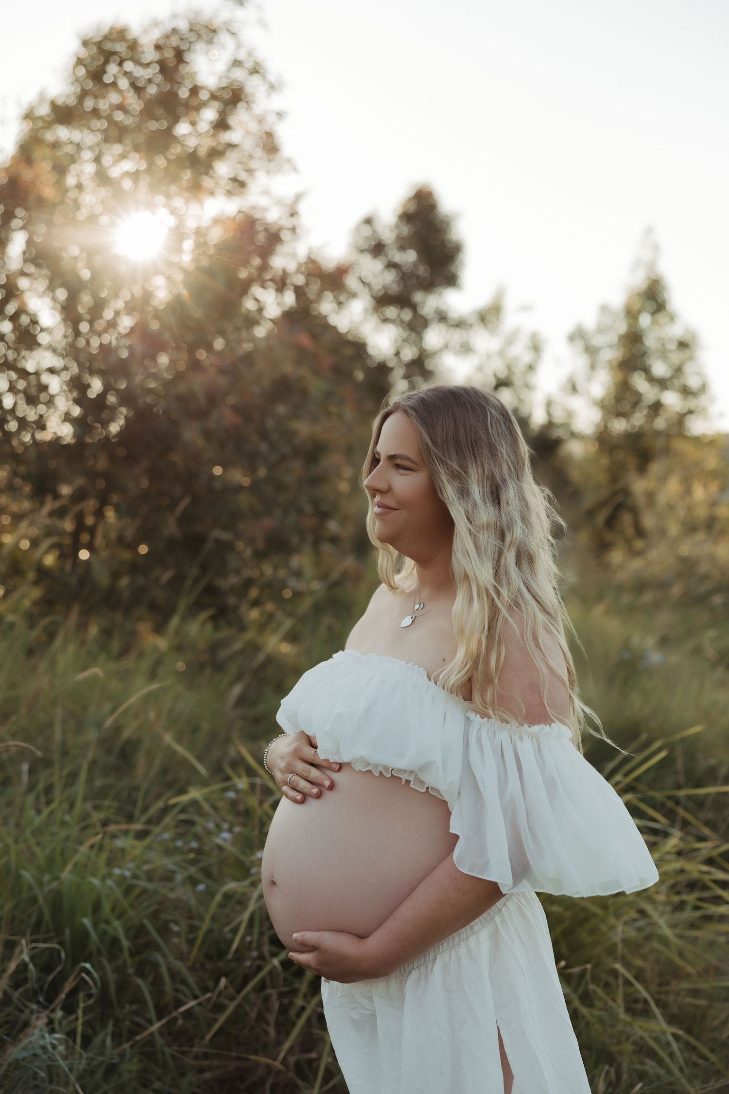 Maternity photo in white dress amid sunlit natural setting.