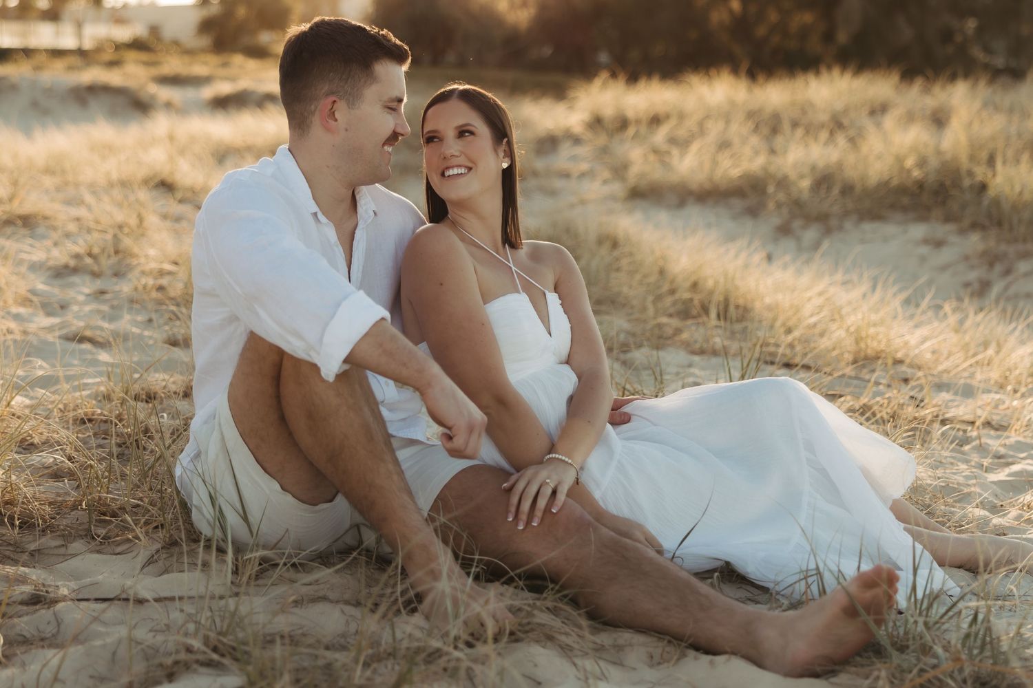 A romantic couple in white outfits reclines on golden grass during a sunset photoshoot.