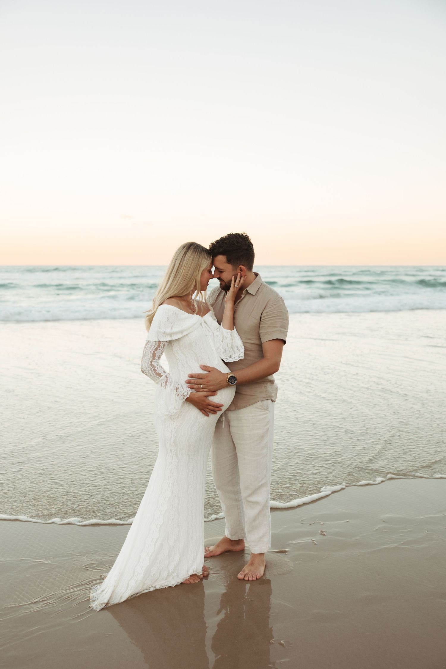 A pregnant couple embraces a Gold Coast beach at sunset with gentle waves rolling in behind them.