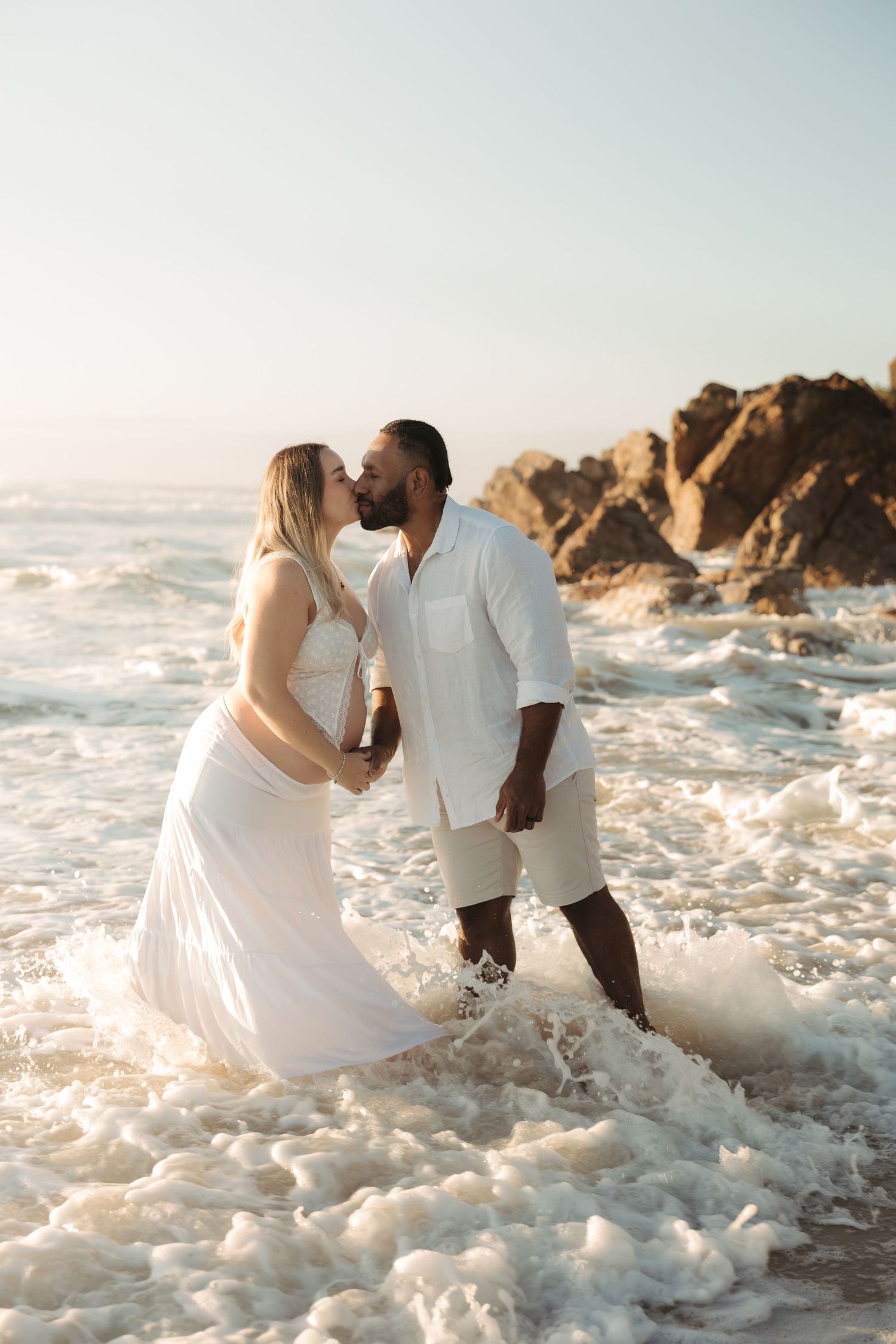A romantic couple in white outfits share a kiss while standing in ocean waves against rocky coastal cliffs at sunset.