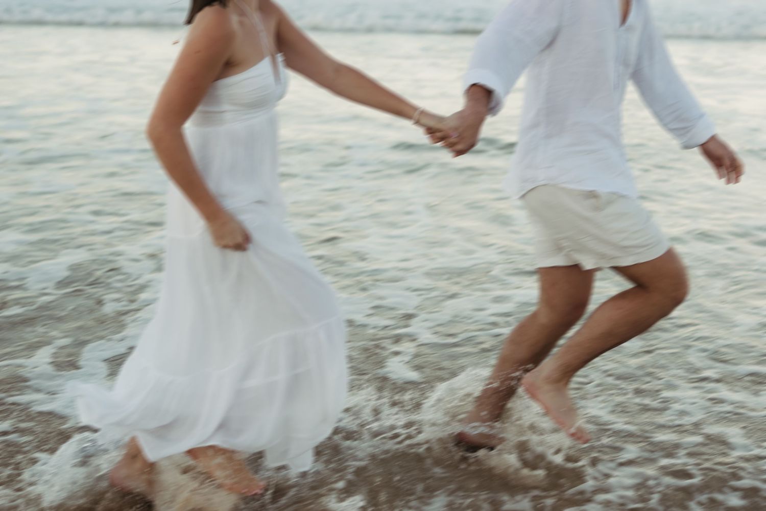 A couple in white clothing holds hands while splashing through ocean waves on a beach at sunset.