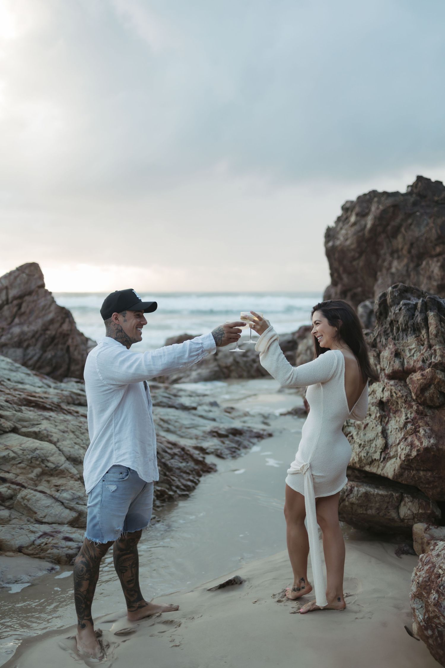 Couple poses playfully on a rocky beach during sunset, creating a series of fun dabbing dance moves together.