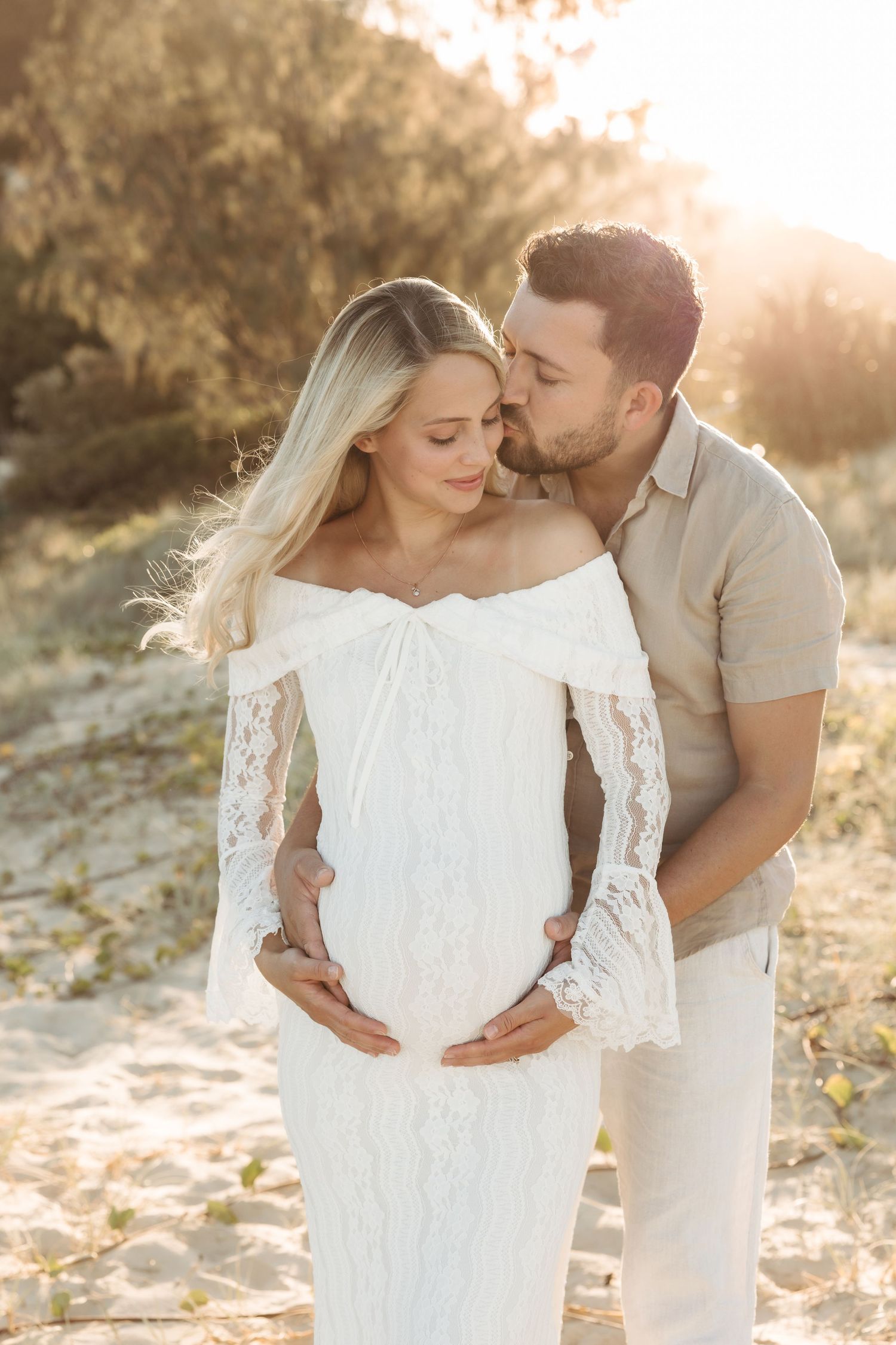 A romantic maternity photo at sunset shows a couple in white and beige embracing on a sandy beach with golden light.