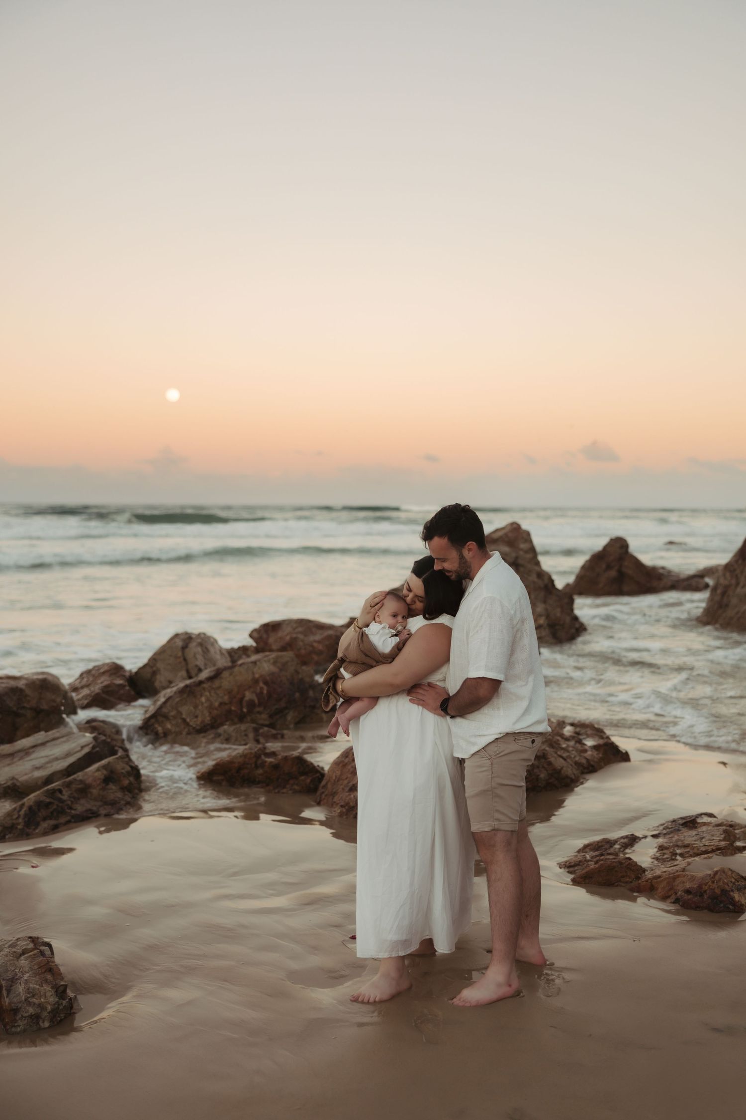 A romantic couple embraces on a rocky beach at sunset with gentle waves and warm orange sky in the background.