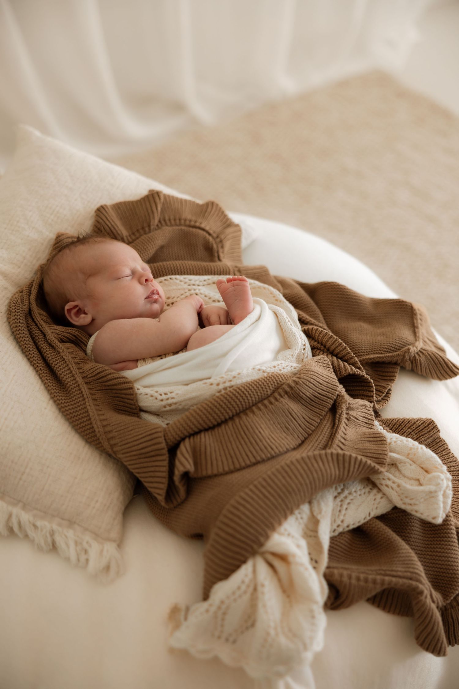 A sleeping newborn baby wrapped in a cozy brown knit blanket and white swaddle on a soft textured surface.