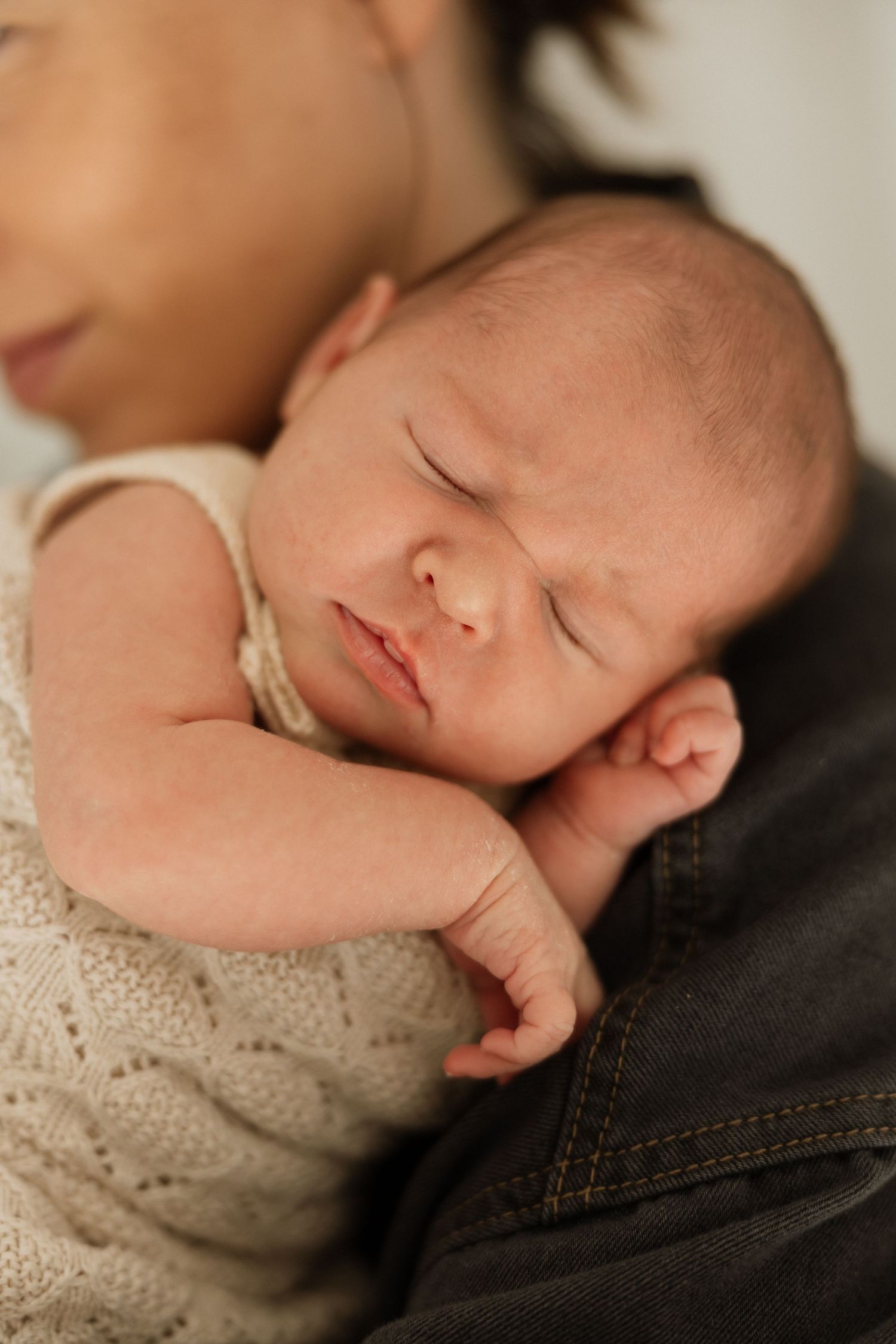 A sleeping newborn baby rests peacefully against a dark background while wearing a cream-colored knit outfit.