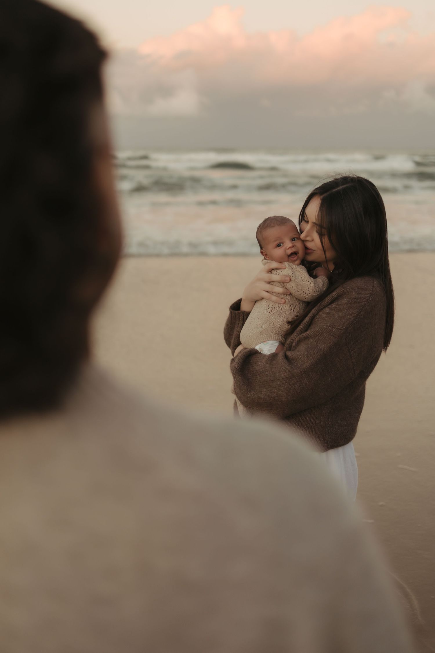 A parent and baby share a tender moment on a misty beach at sunset, wrapped in a cozy brown sweater.