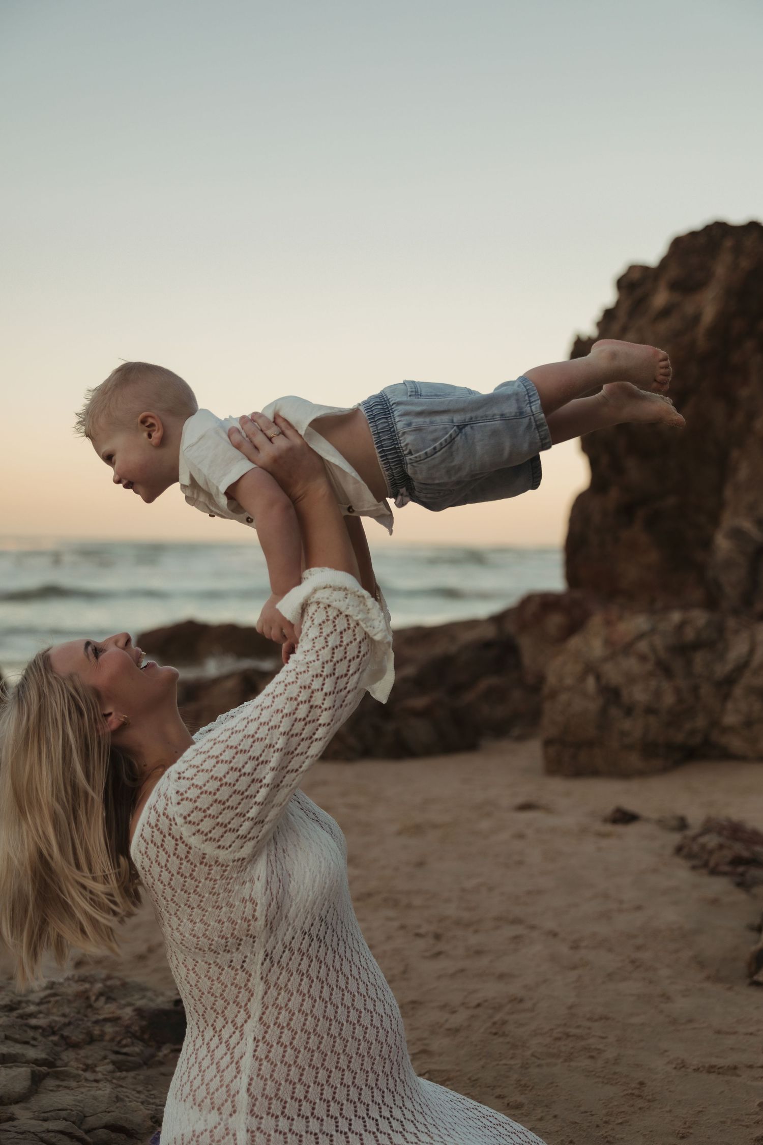A tender moment between parent and child playing on a beach at sunset with rocky cliffs in the background.