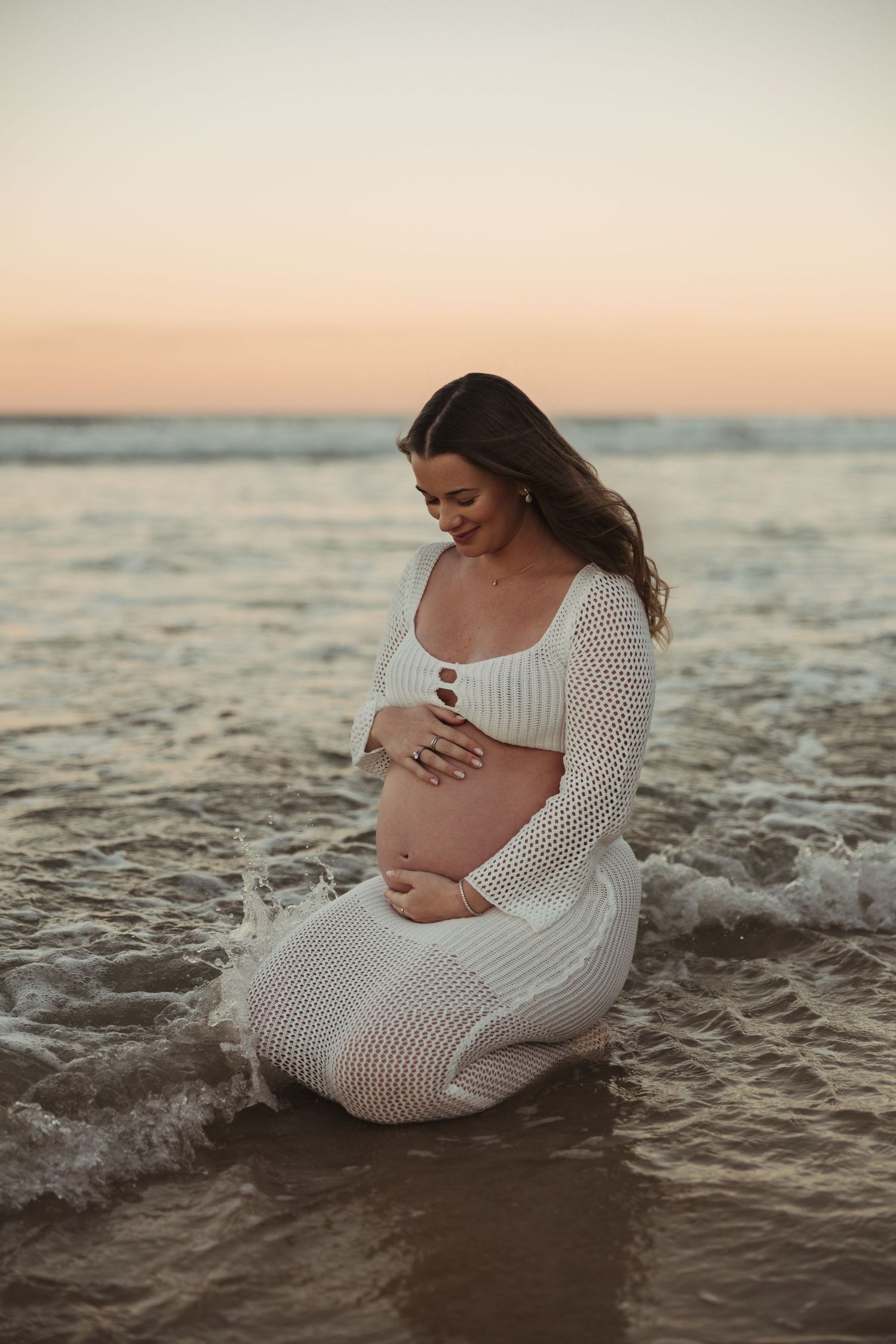 Expectant mother in white crochet dress sitting in ocean waves during sunset maternity photoshoot.